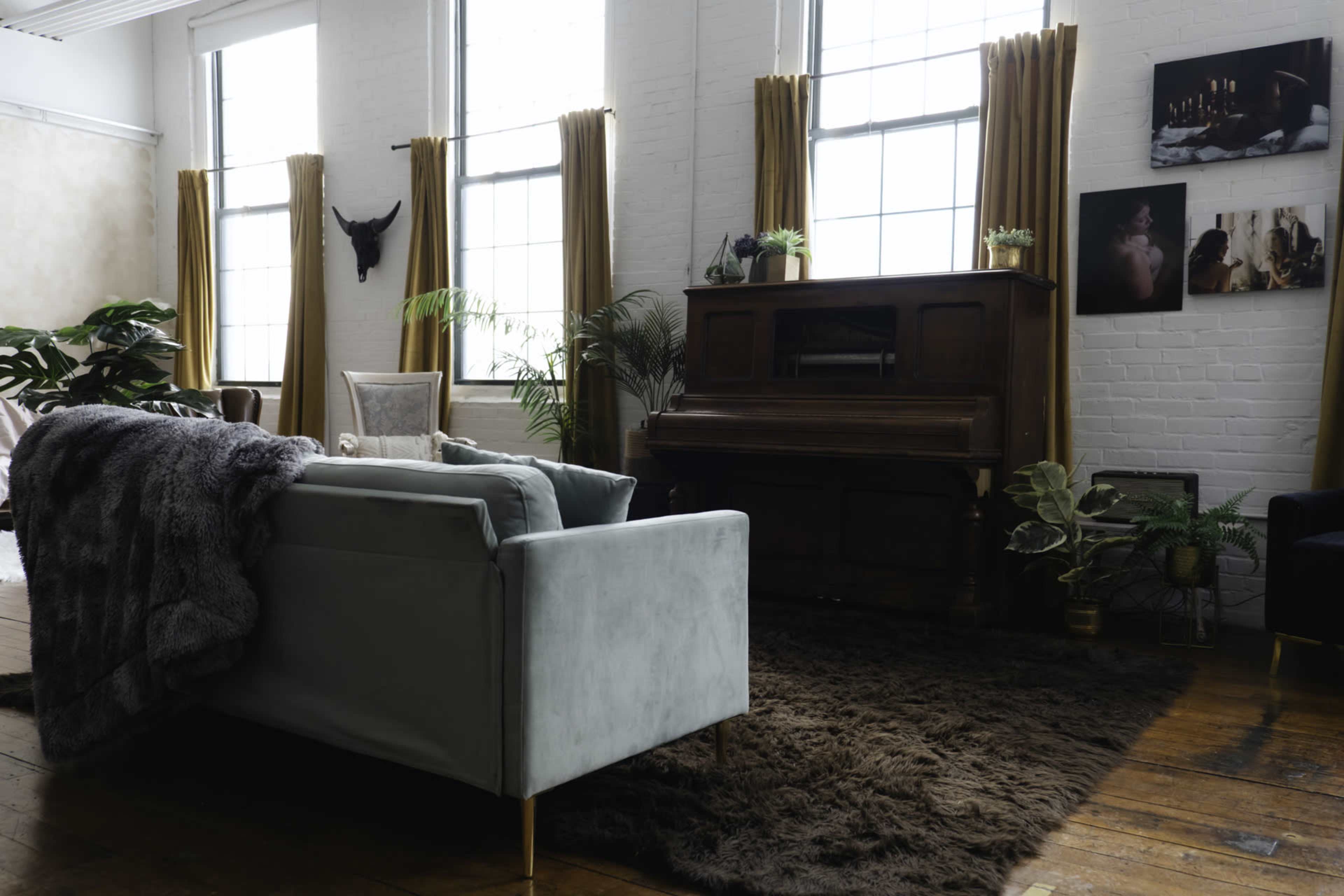 A modern living room features a light blue sofa with a textured throw, a wooden piano, large windows with golden curtains, and various plants and artwork on the walls.