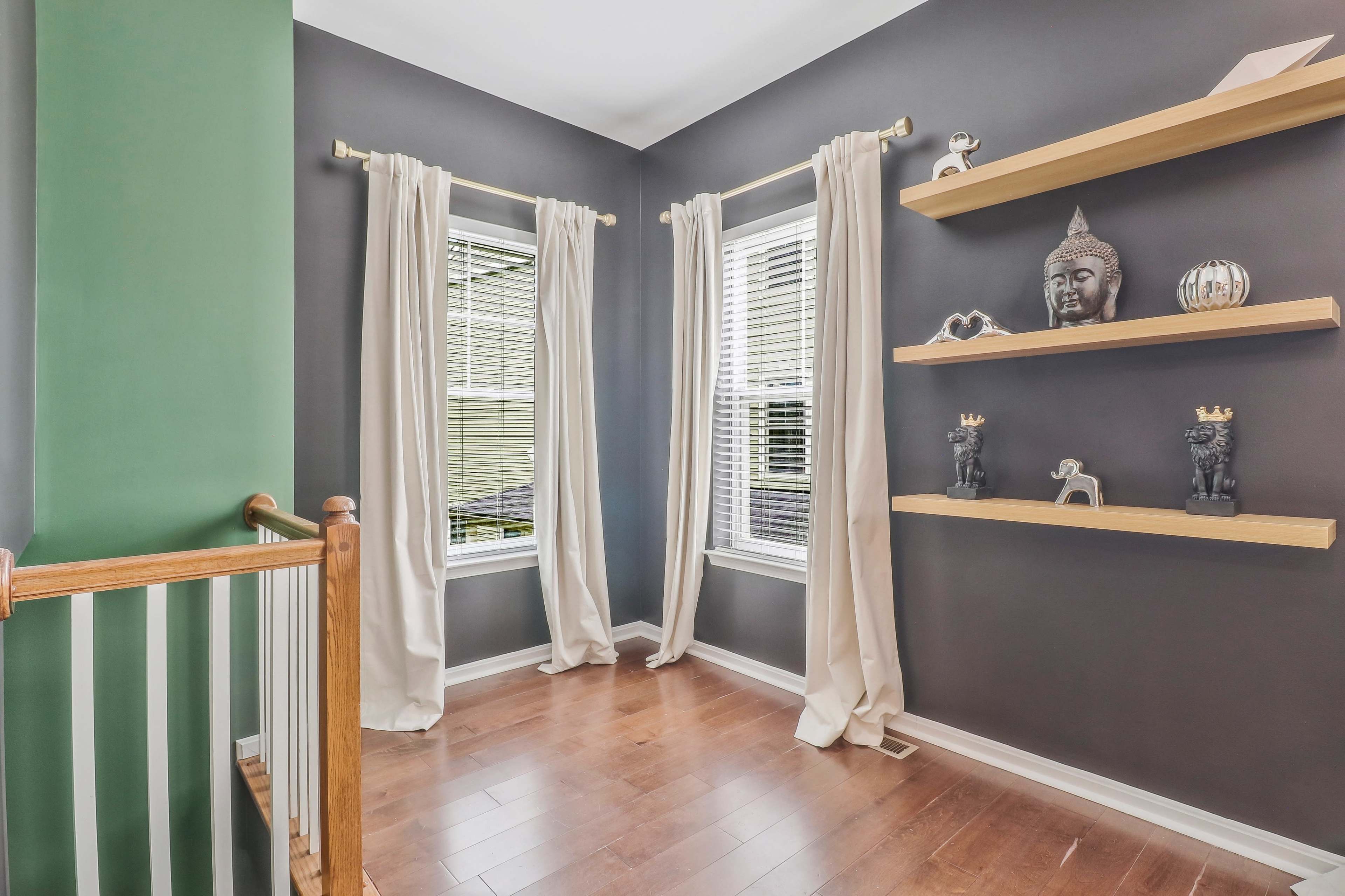 The room features two windows with light-colored curtains, a green accent wall, hardwood flooring, and wooden shelves displaying decorative items.