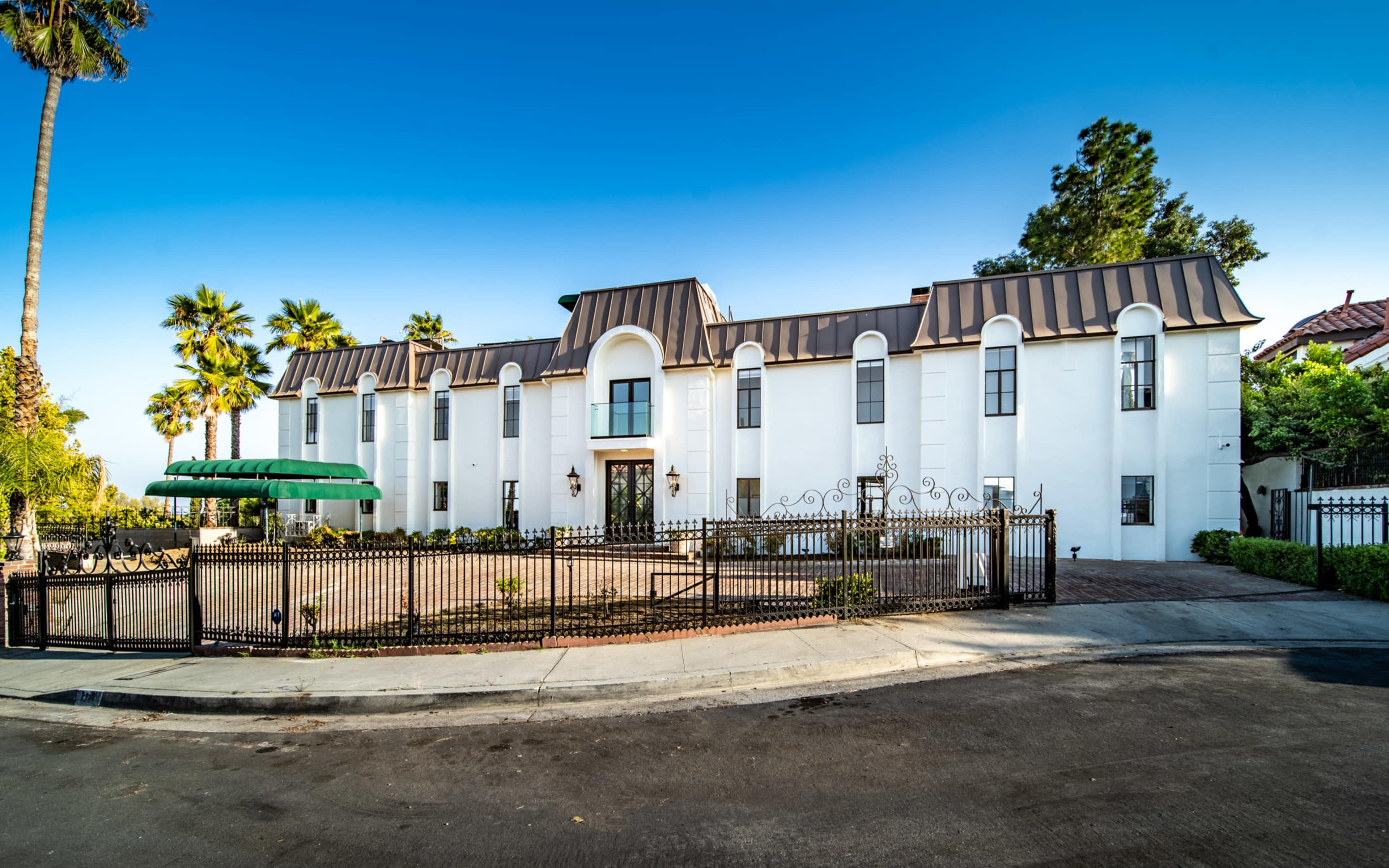 The image shows a large, white multi-story building with a brown metal roof, surrounded by a black wrought-iron fence and palm trees.