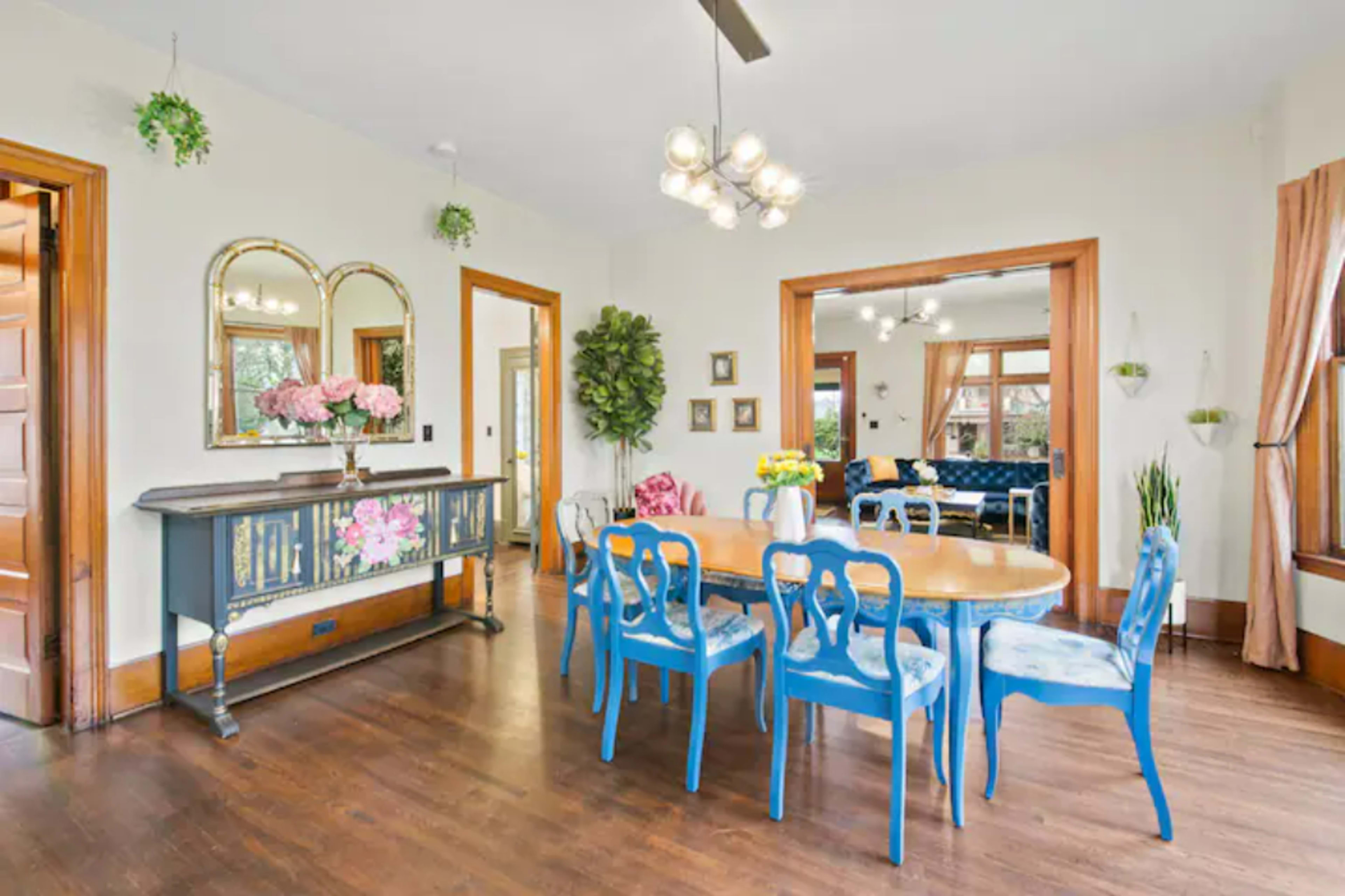A well-lit dining area with a round wooden table surrounded by blue chairs, a sideboard with floral decorations, and views into a living room through an open archway.