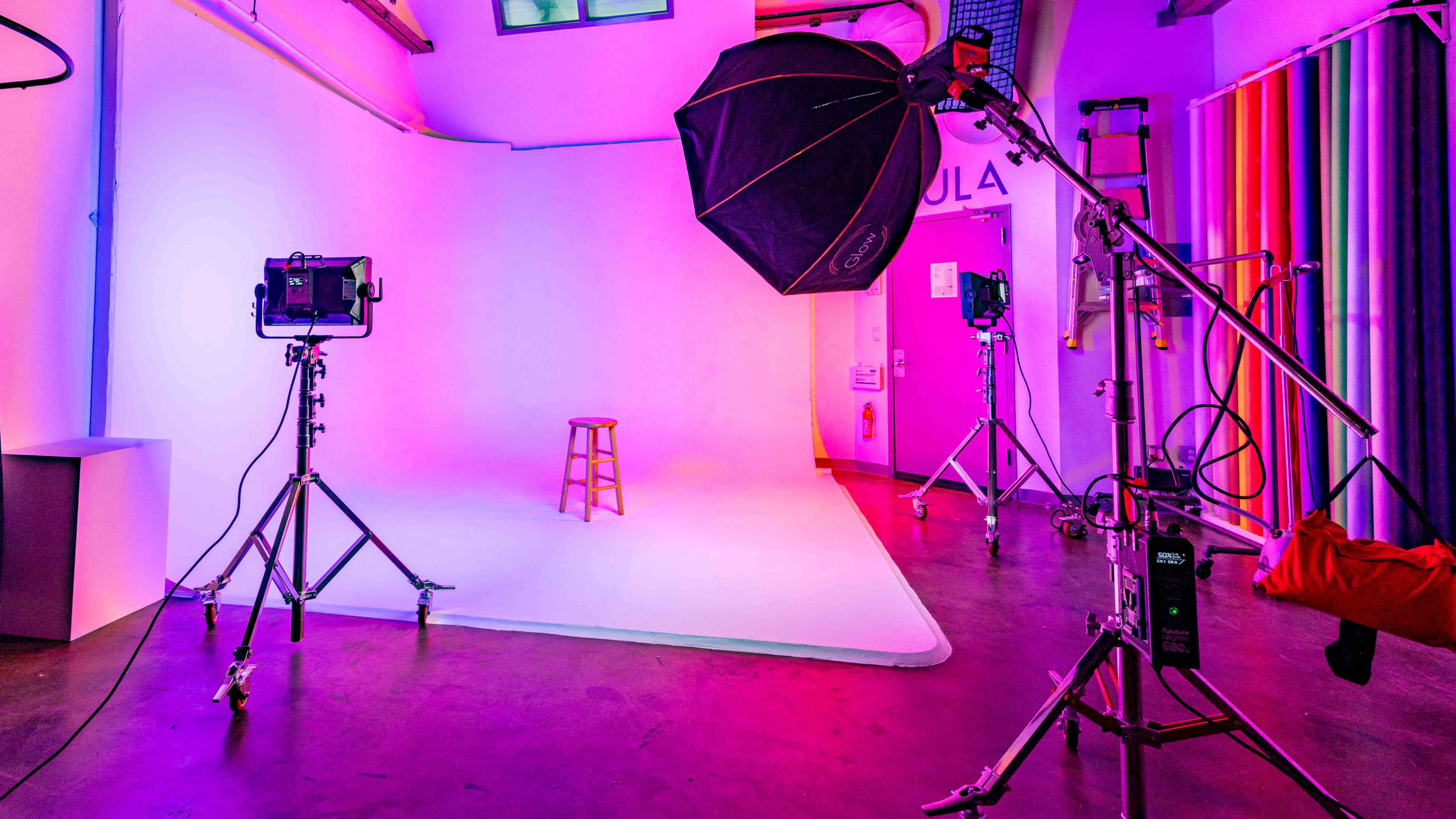 A photography studio is set up with a stool in the center, surrounded by various lighting equipment and colorful backdrops.