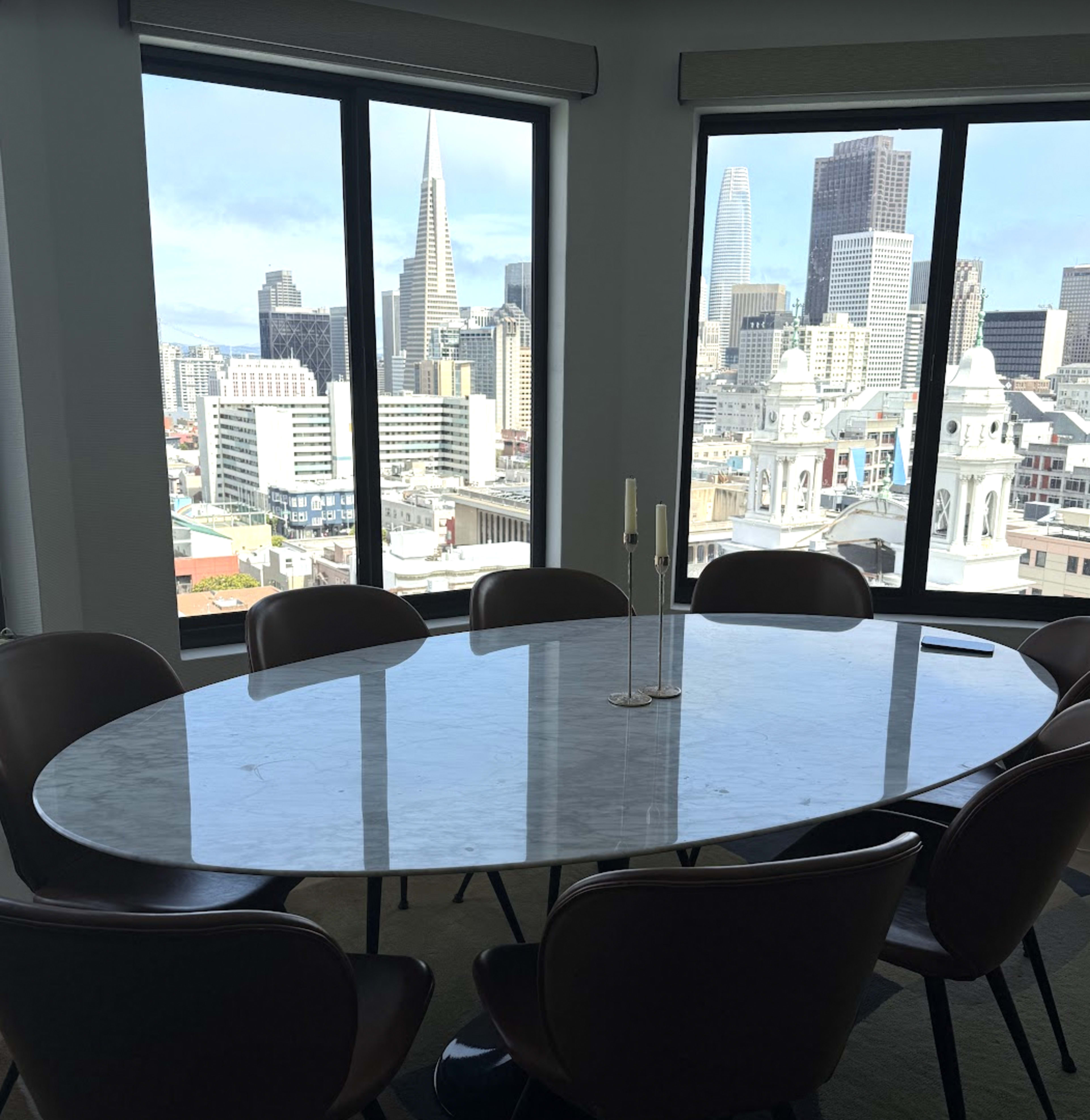 A conference room with an oval marble table and chairs overlooks a skyline featuring prominent high-rise buildings and the Transamerica Pyramid in the background.