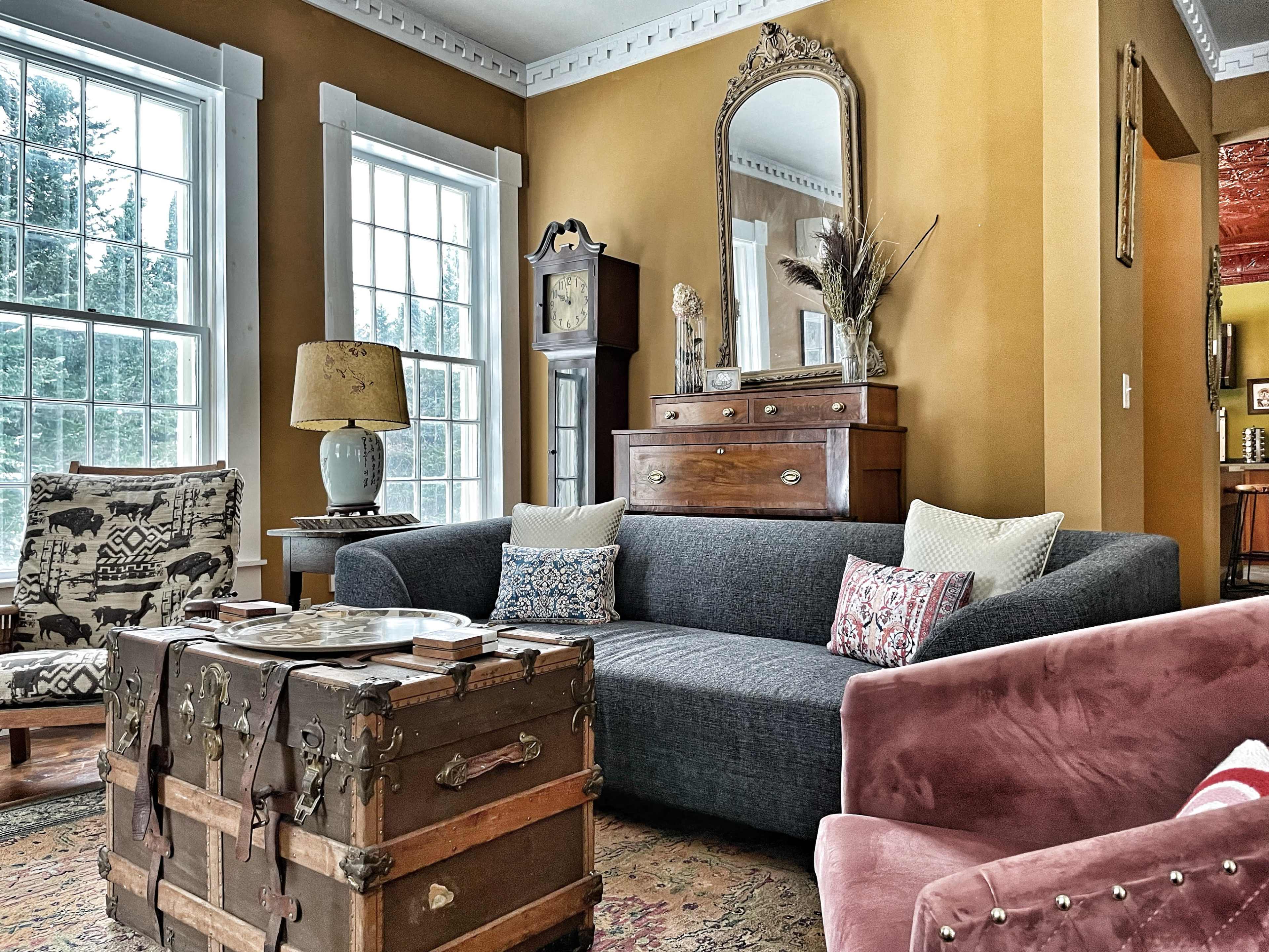 The living room features a gray sofa with decorative pillows, a vintage trunk table, a wooden dresser with a mirror, and large windows allowing natural light to fill the space.