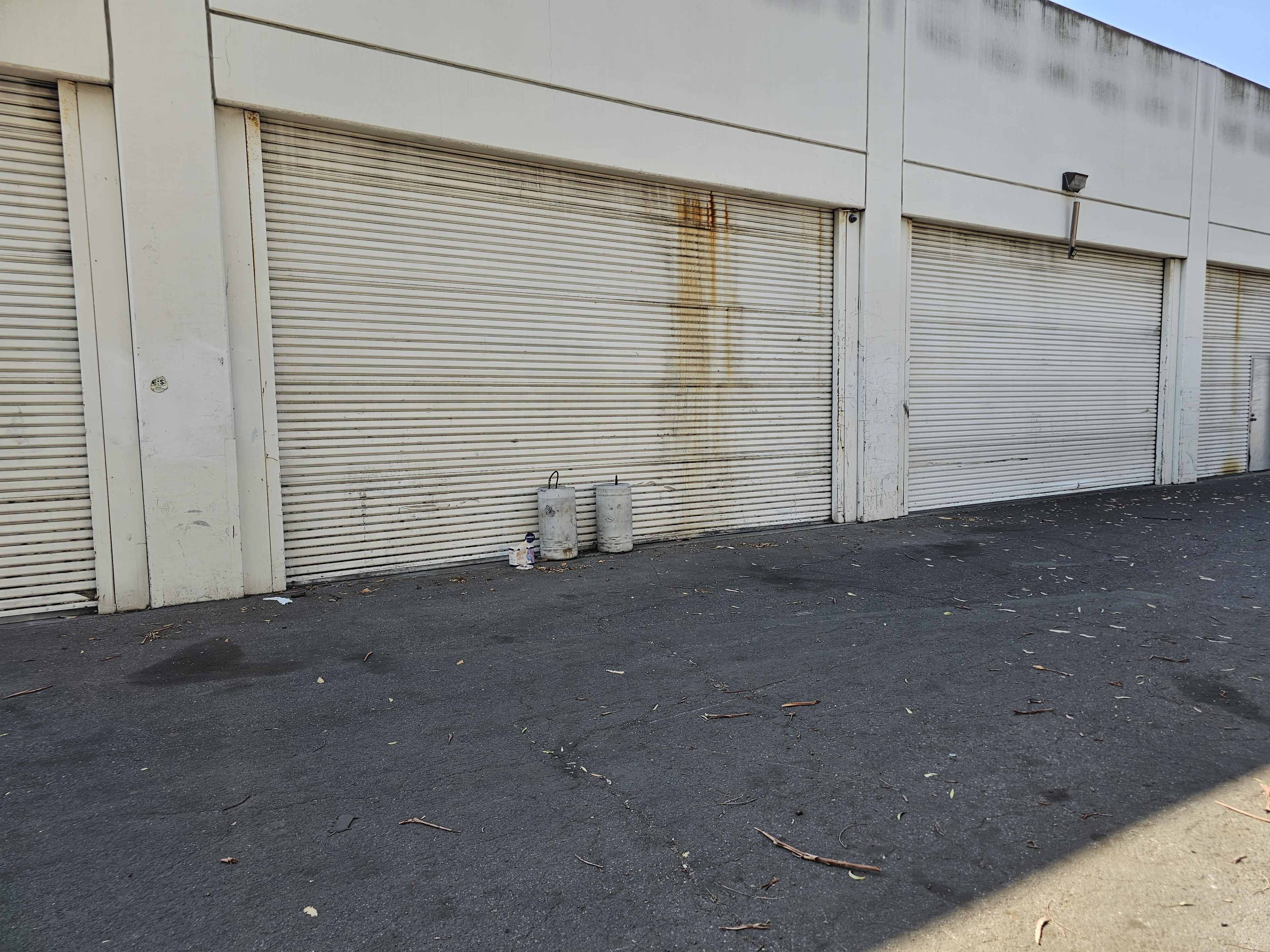 The image shows a row of closed metal roller doors with a stained wall beside a paved, litter-strewn area.