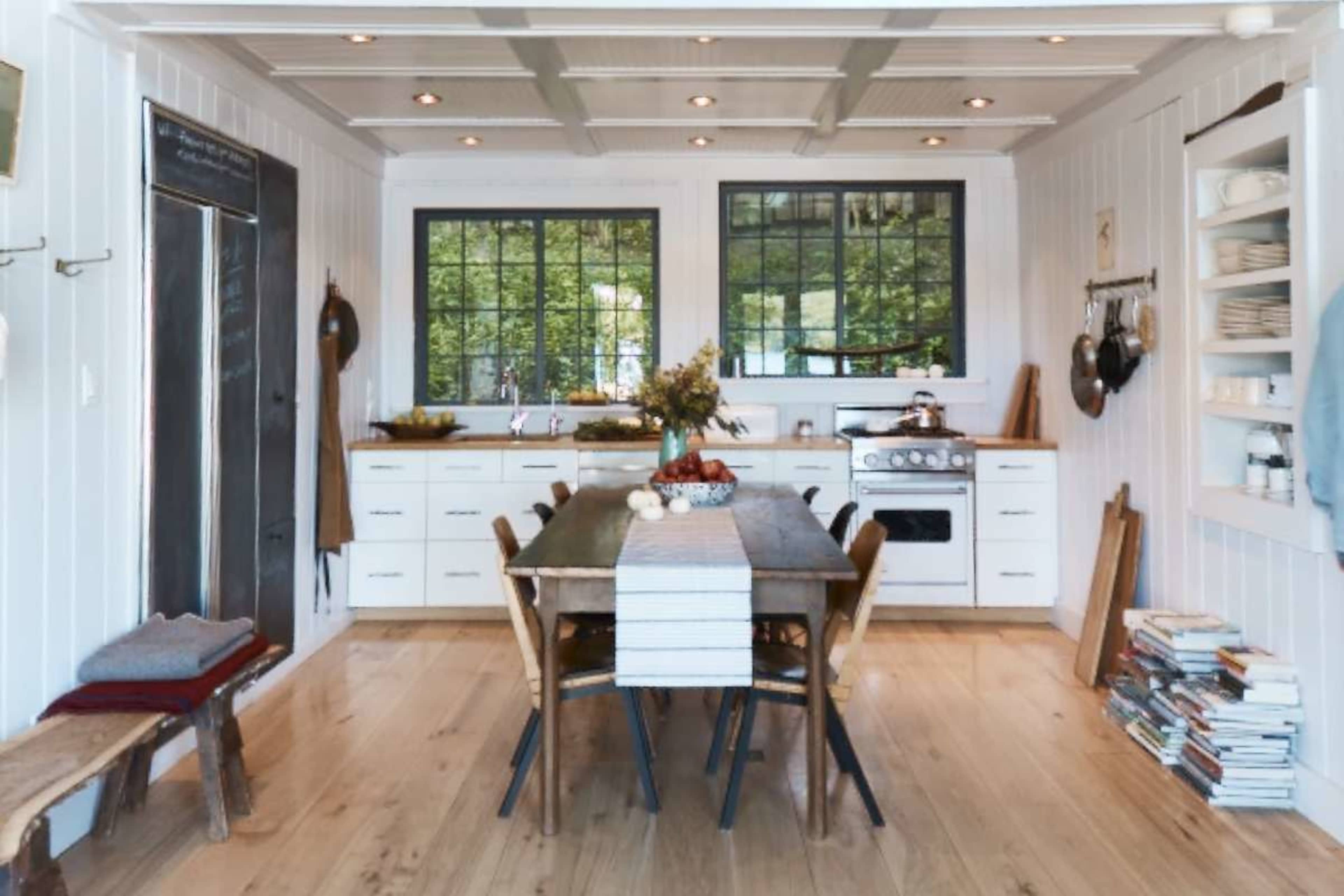 The image shows a bright kitchen with white walls, a wooden dining table surrounded by chairs, and large windows letting in natural light.