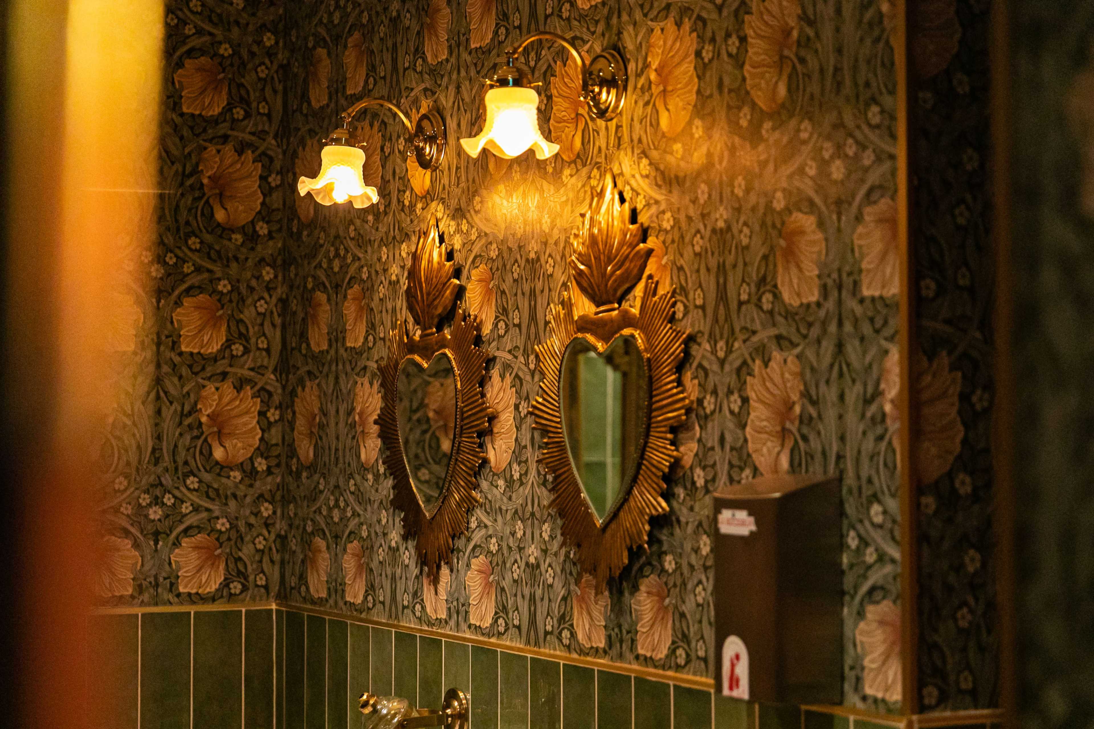 A bathroom featuring floral wallpaper, two heart-shaped mirrors, and vintage light fixtures.