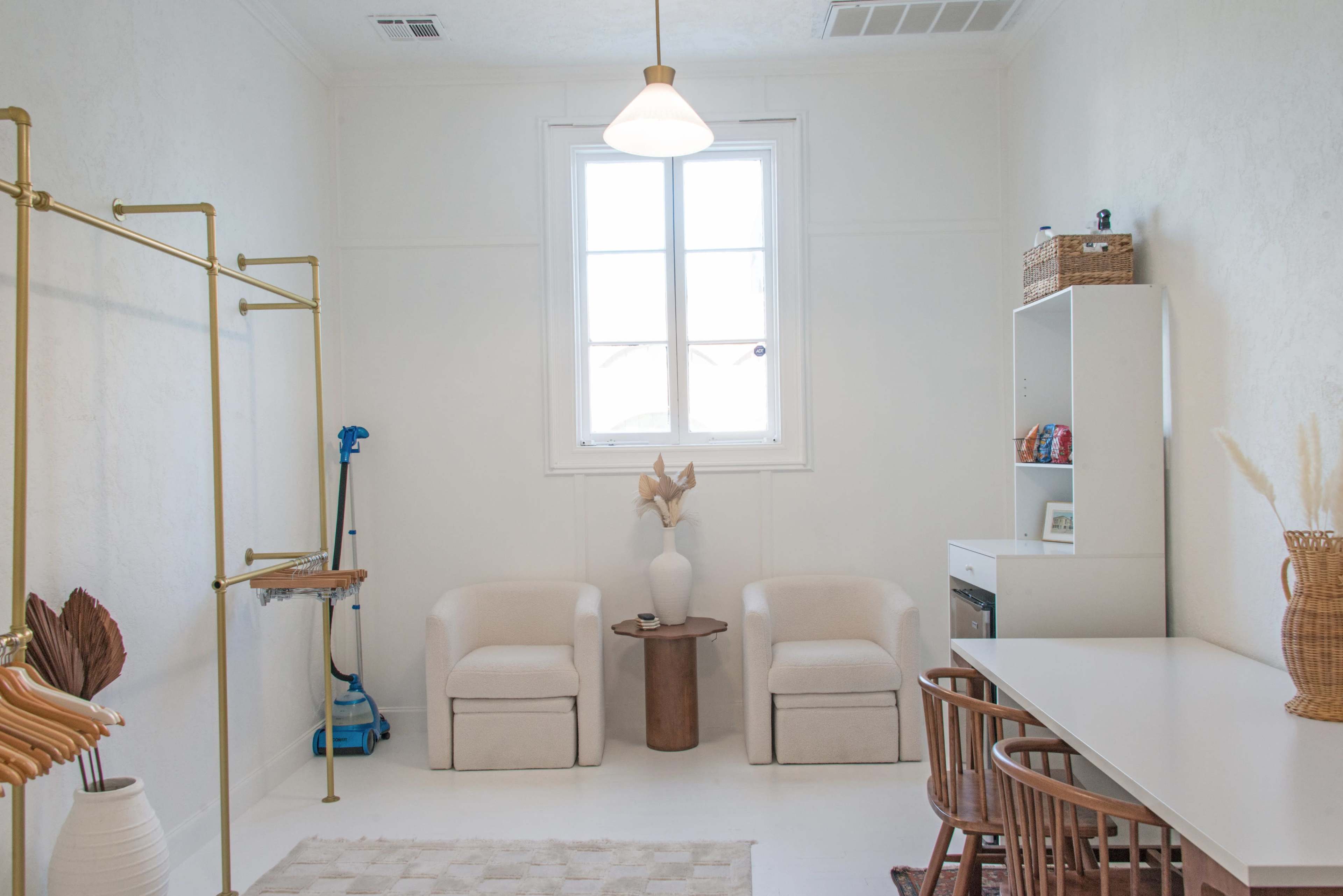 The image shows a bright, minimalist room featuring two chairs, a small table, a desk with chairs, and a window allowing natural light to enter.