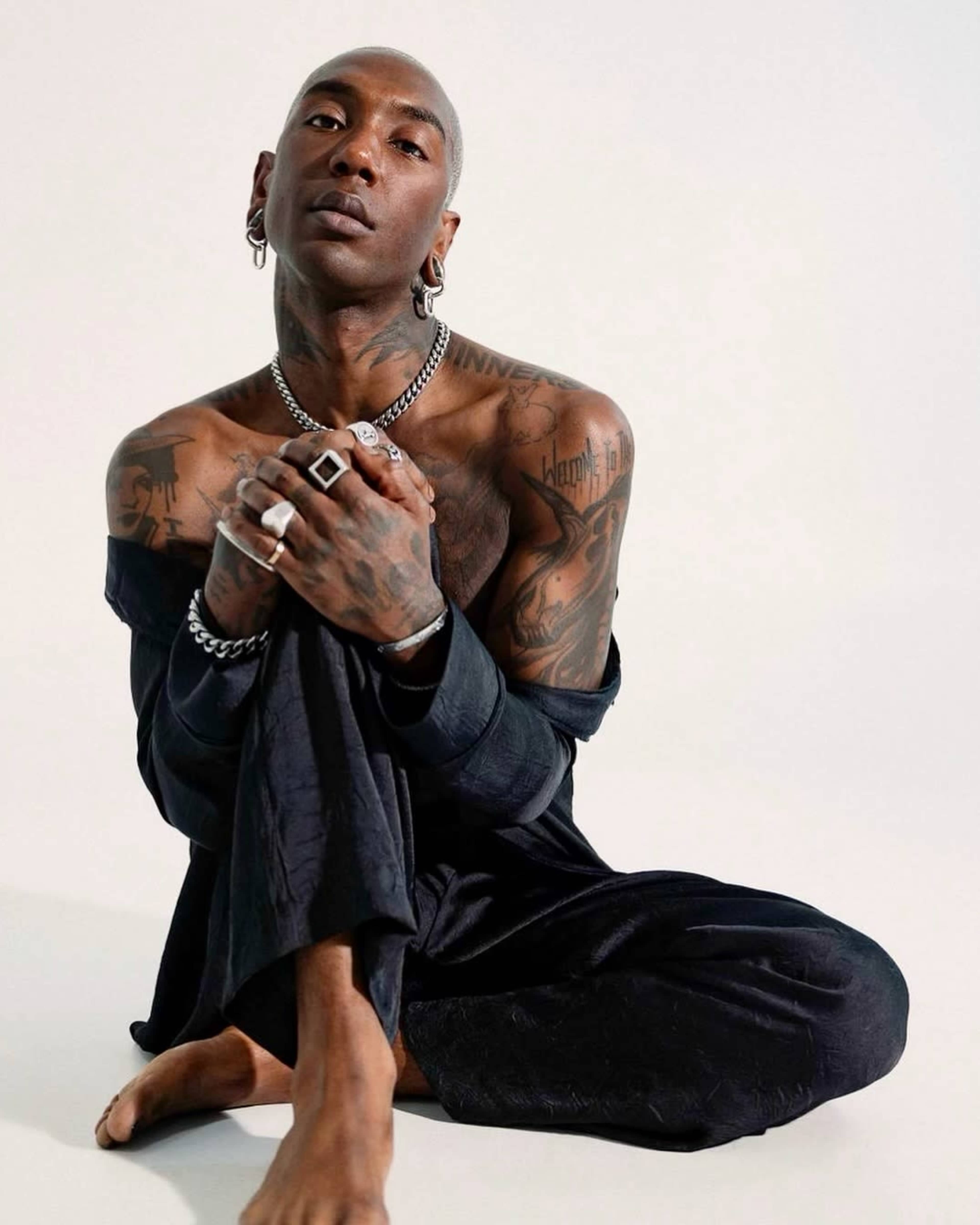 A person with tattoos sits on the floor, dressed in dark clothing with an off-shoulder top, posing with their knees drawn up.