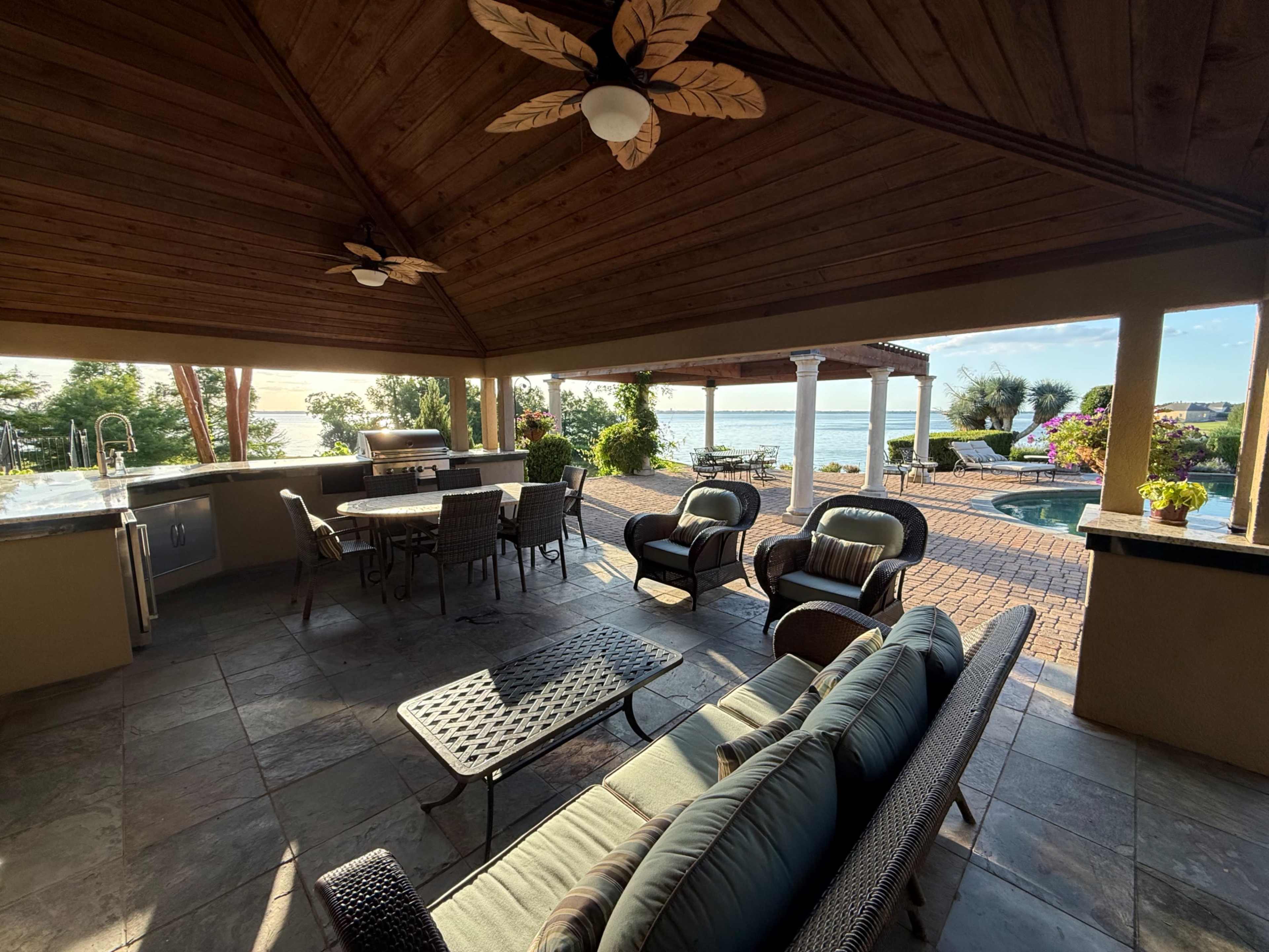 The image shows a covered outdoor patio area with seating, a dining table, and a view of a swimming pool and waterfront landscape.