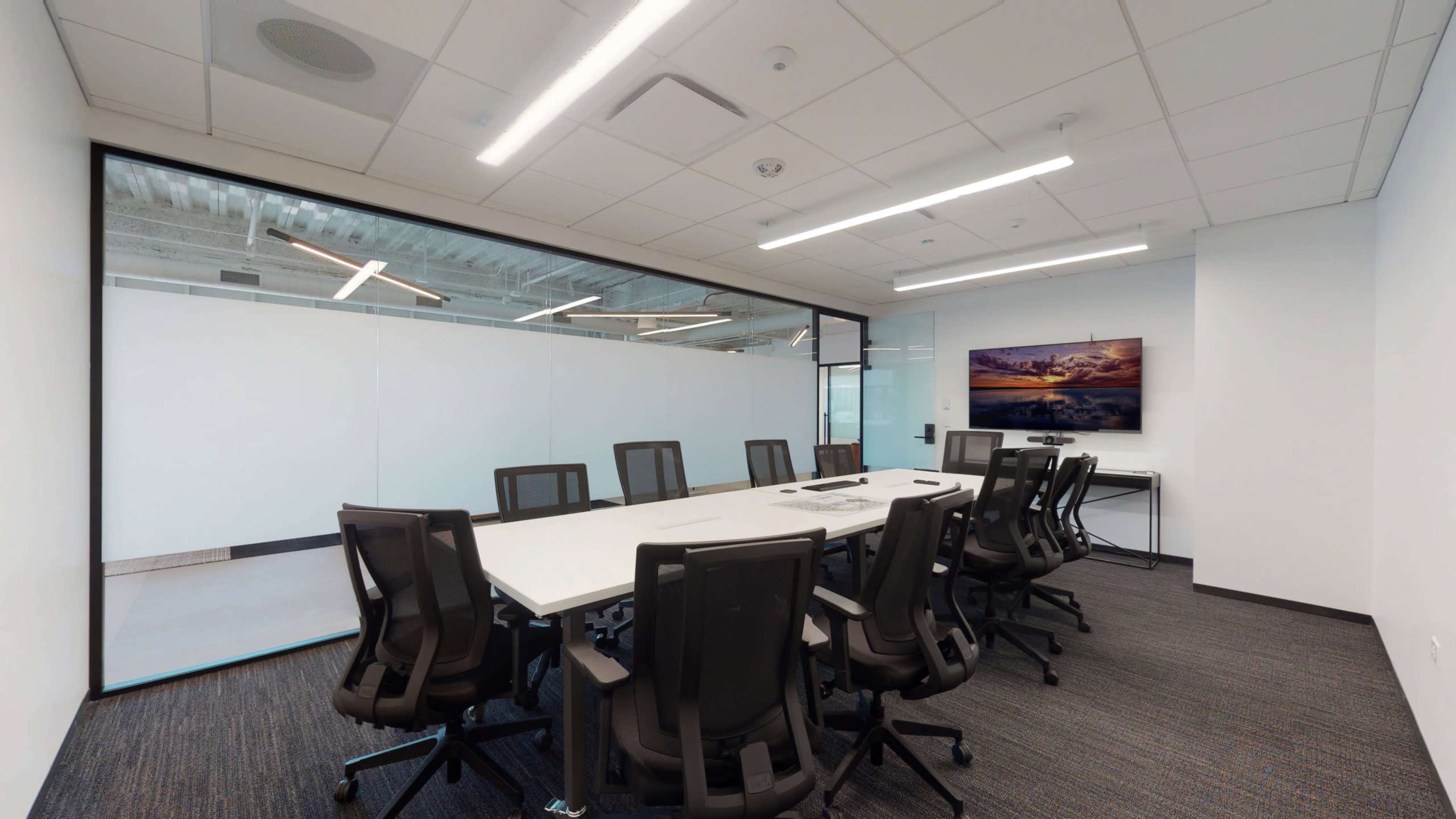 A conference room features a long white table surrounded by black chairs, with glass walls and a large landscape artwork on display.