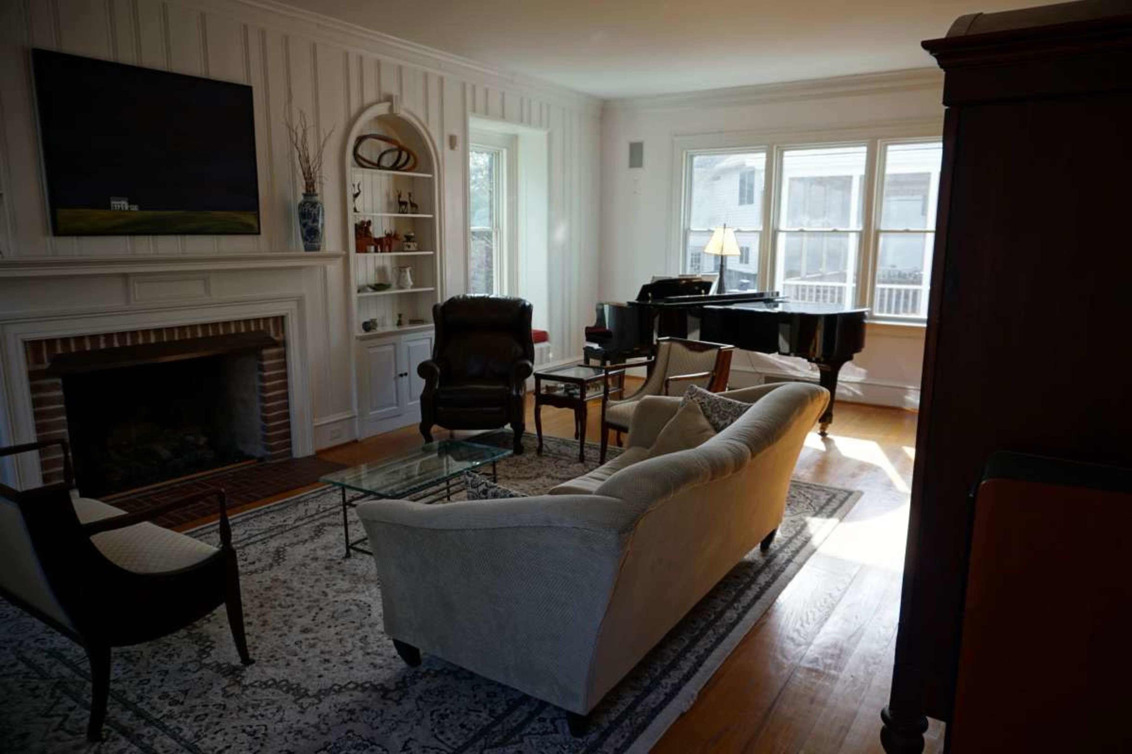 The living room features a fireplace, a grand piano, a beige sofa, and multiple large windows allowing natural light to fill the space.