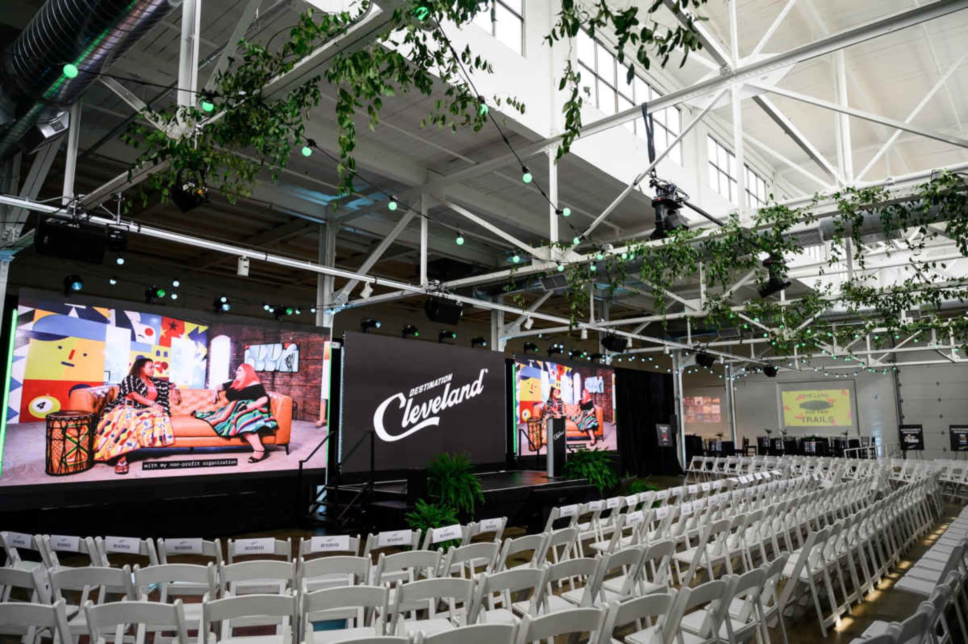 The image shows a spacious event venue with rows of white chairs facing a large screen displaying content, surrounded by decorative greenery and ample lighting.