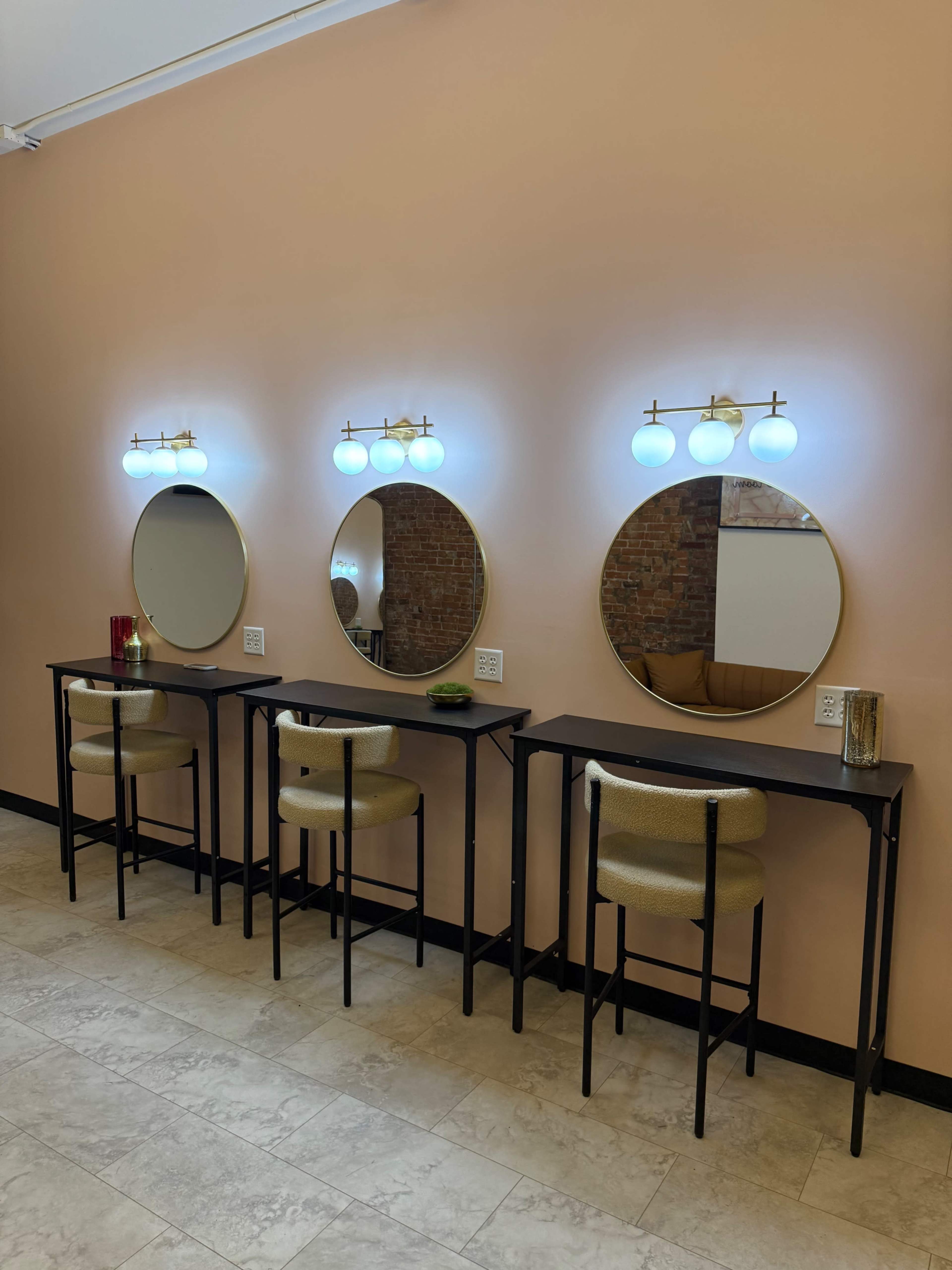 The image shows a wall with three circular mirrors illuminated by light fixtures above, paired with three high stools at sleek, narrow tables against a pastel-colored backdrop.