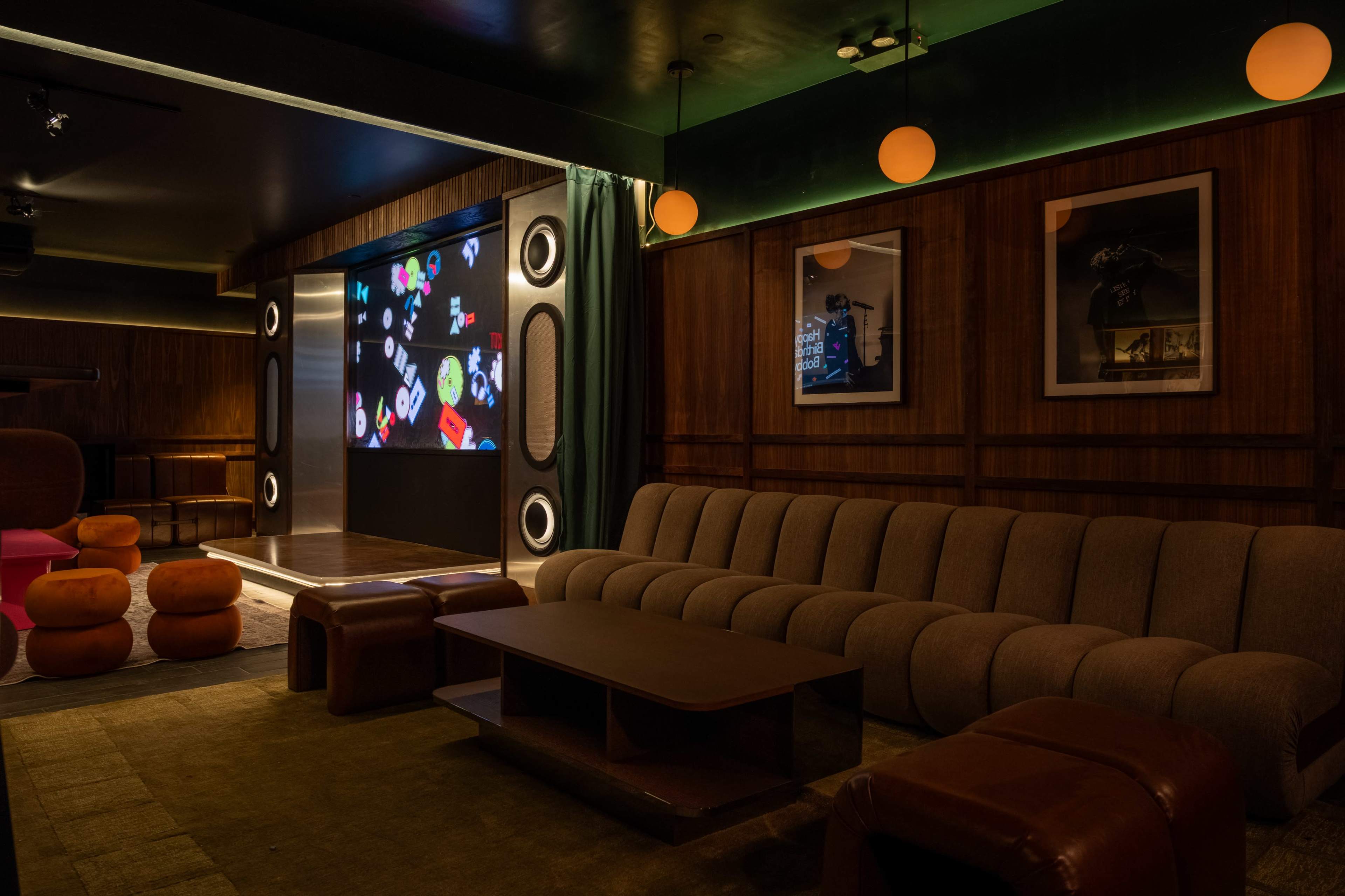 The image shows a dimly lit lounge area with a large screen displaying colorful graphics, a brown sectional couch, and various orange poufs arranged around a wooden coffee table.
