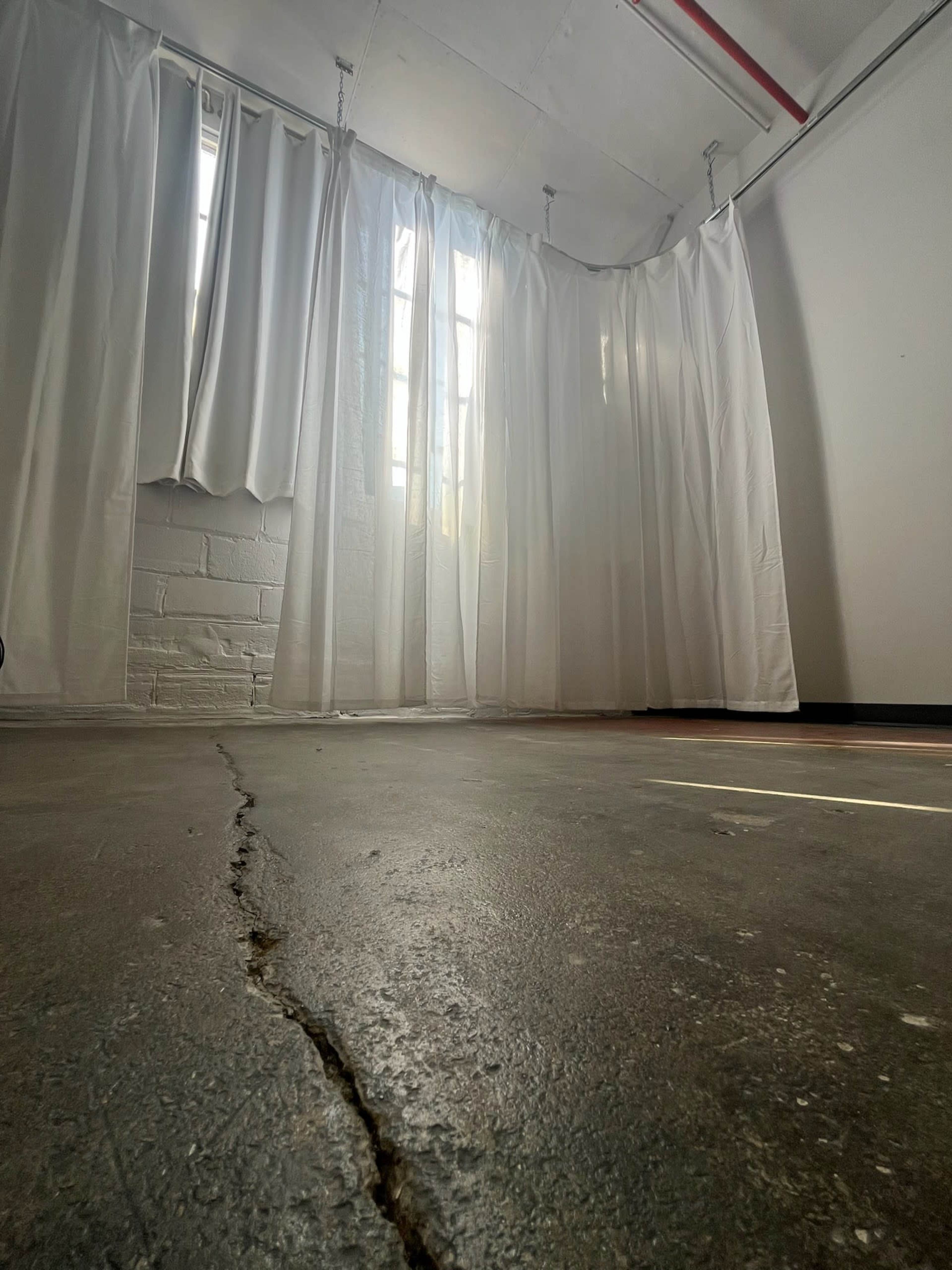 The image shows a room with a cracked concrete floor and white curtains near windows, allowing light to enter.