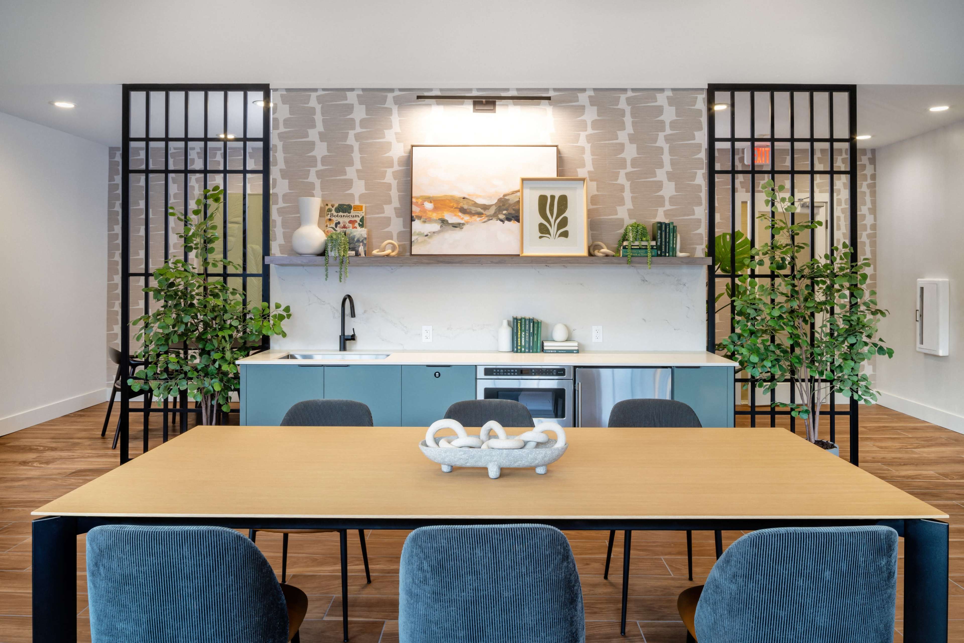 The image shows a modern common area featuring a dining table surrounded by chairs, a kitchen counter with appliances, and decorative plants on either side of a patterned wall.