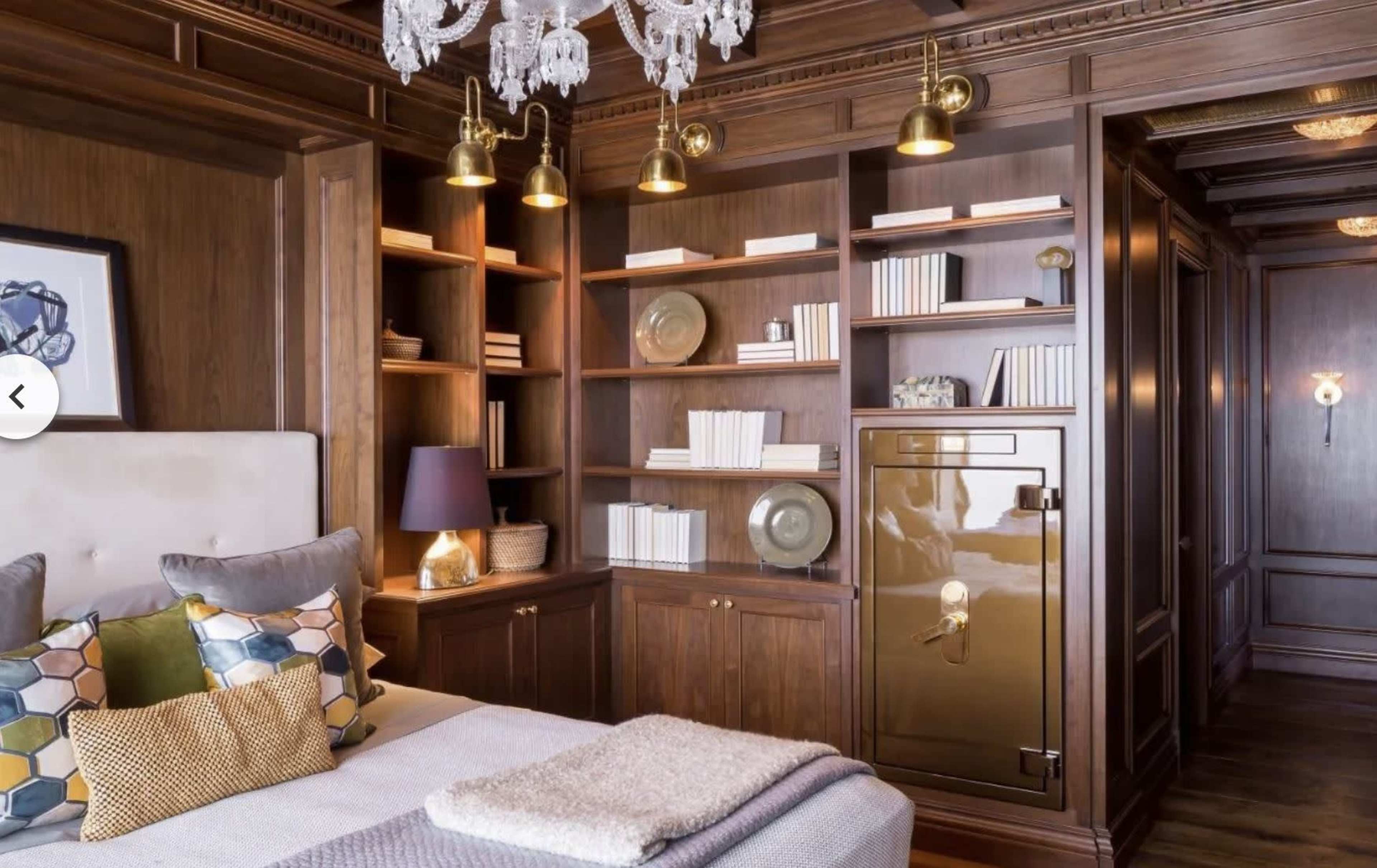 A well-designed bedroom featuring a wooden bookshelf filled with books and decorative items, a bed with layered pillows, and a gold safe integrated into the cabinetry.