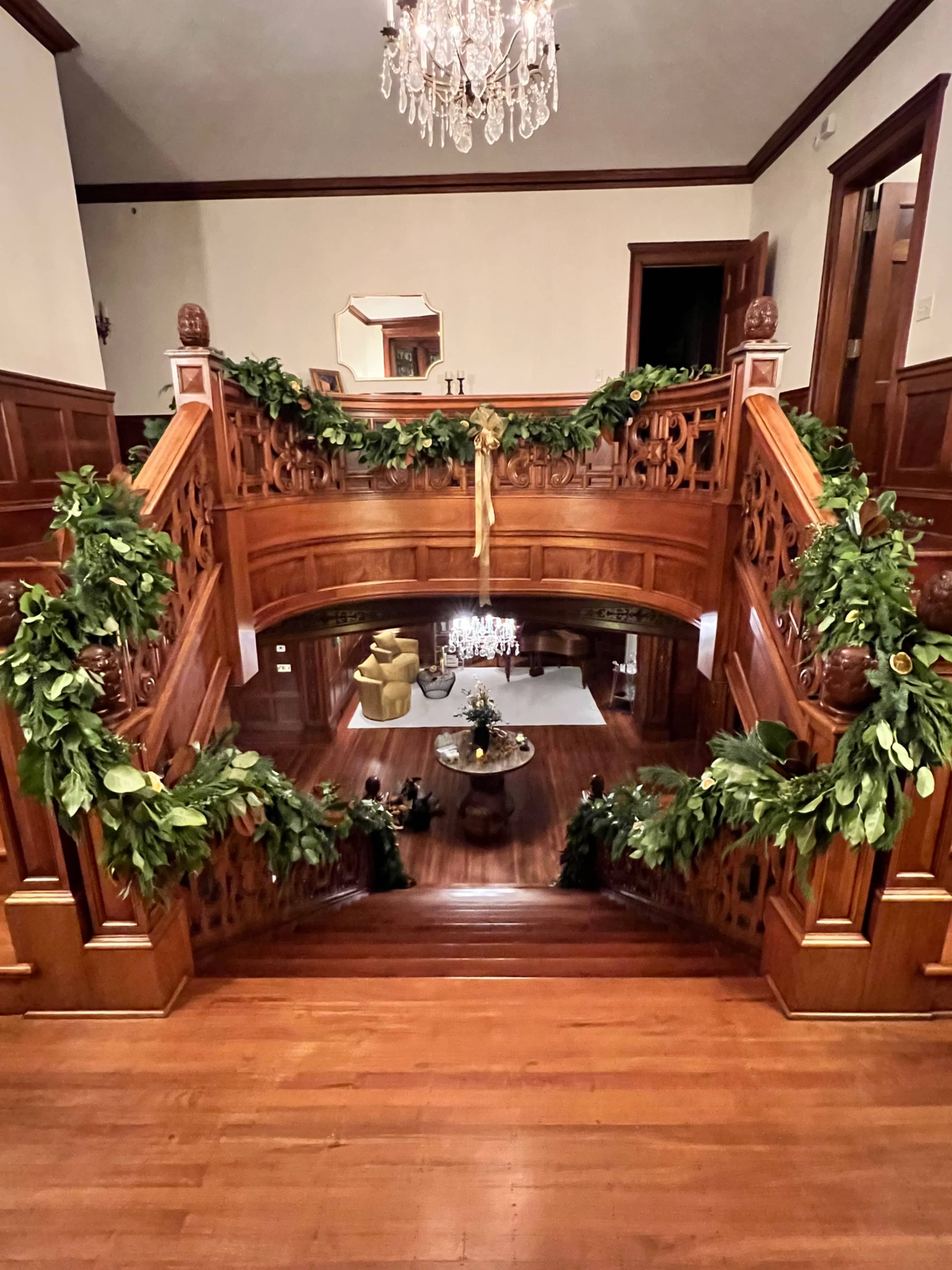 The image shows a grand staircase with wooden railings adorned with green garlands, leading down to a spacious living area with decorative furnishings.