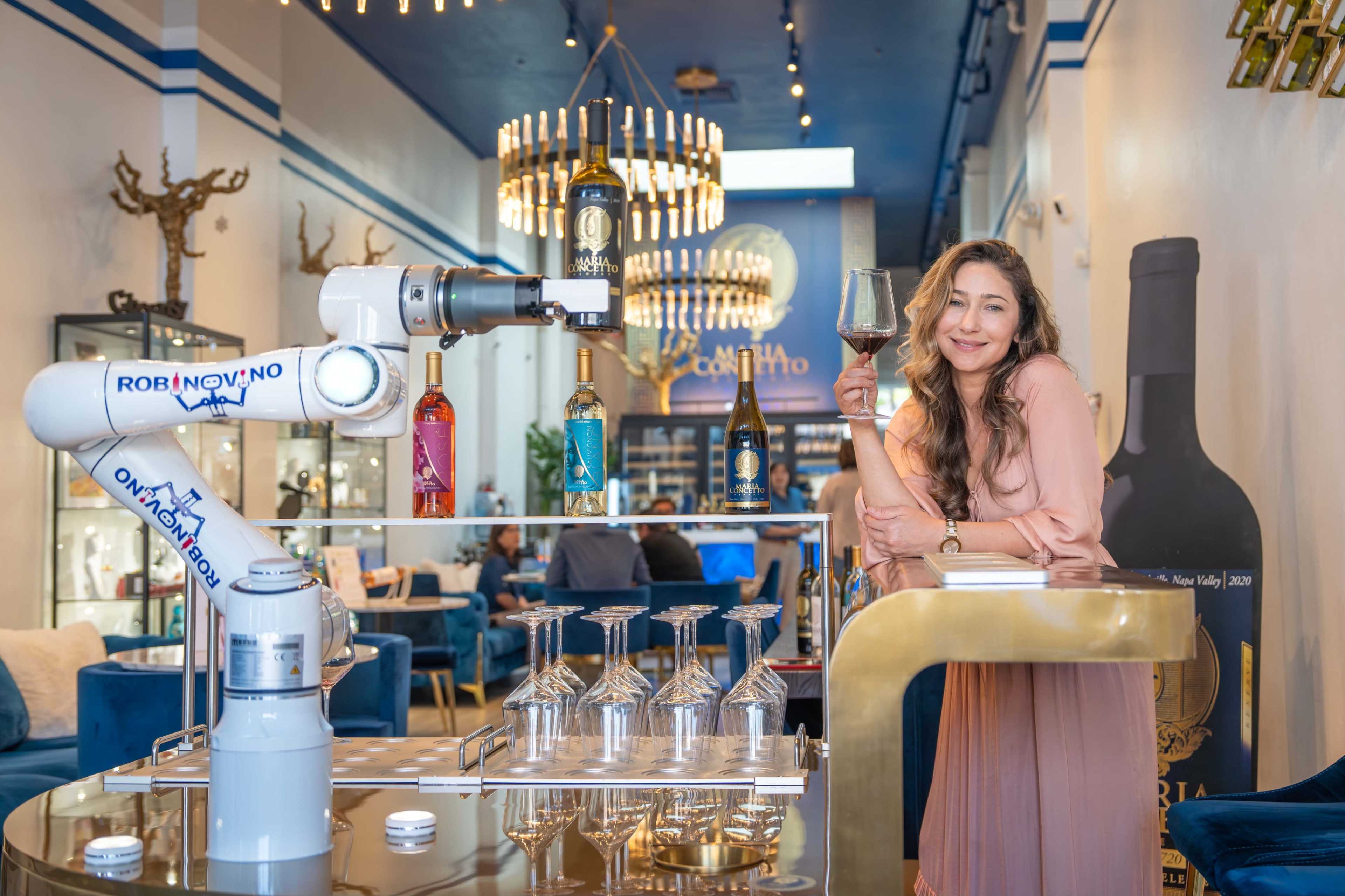A robotic arm is serving drinks while a woman stands next to a table in a modern bar filled with stylish furniture and decorative elements.