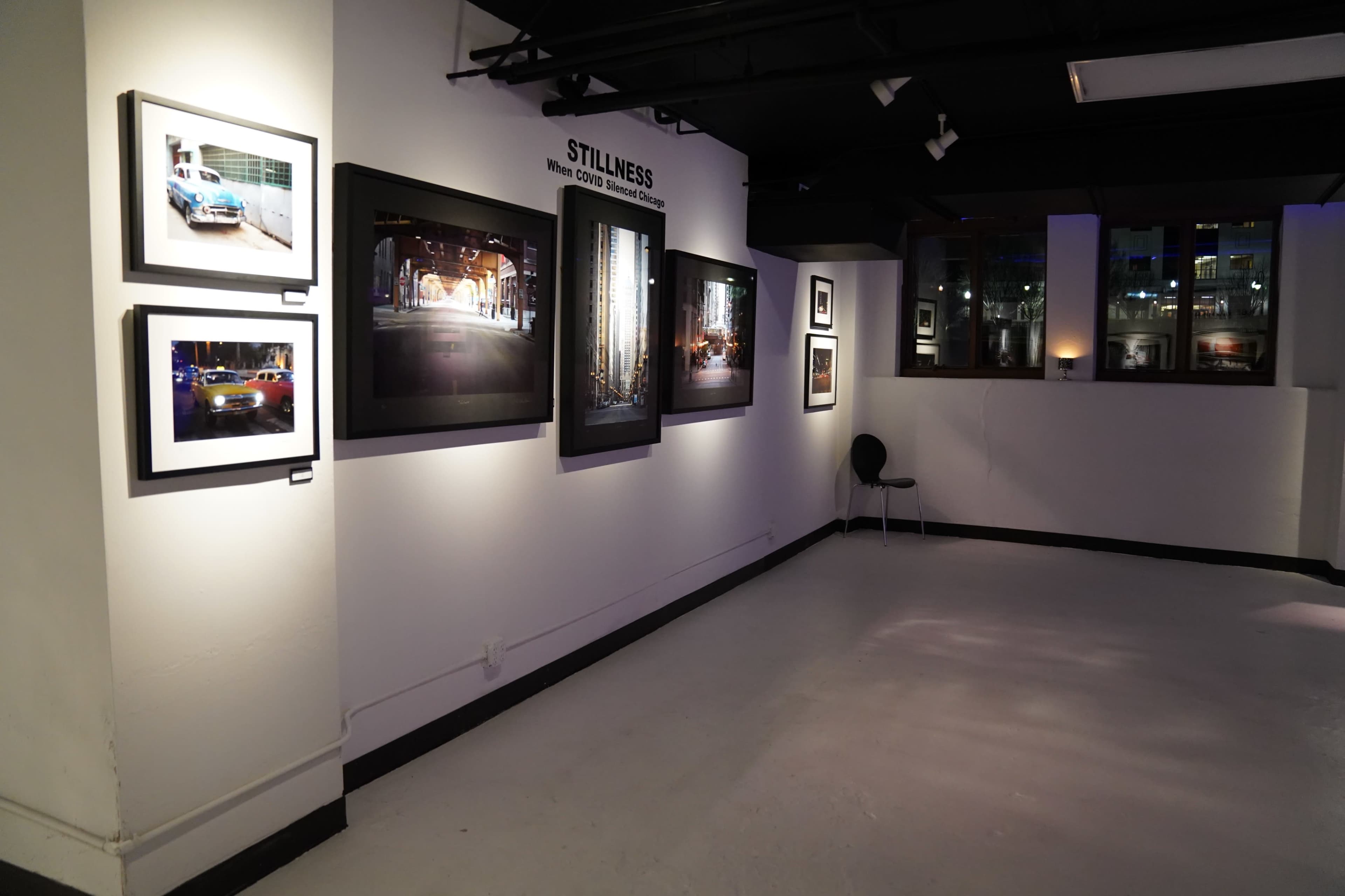 A gallery space with photographs displayed on the walls and a single black chair in the corner.