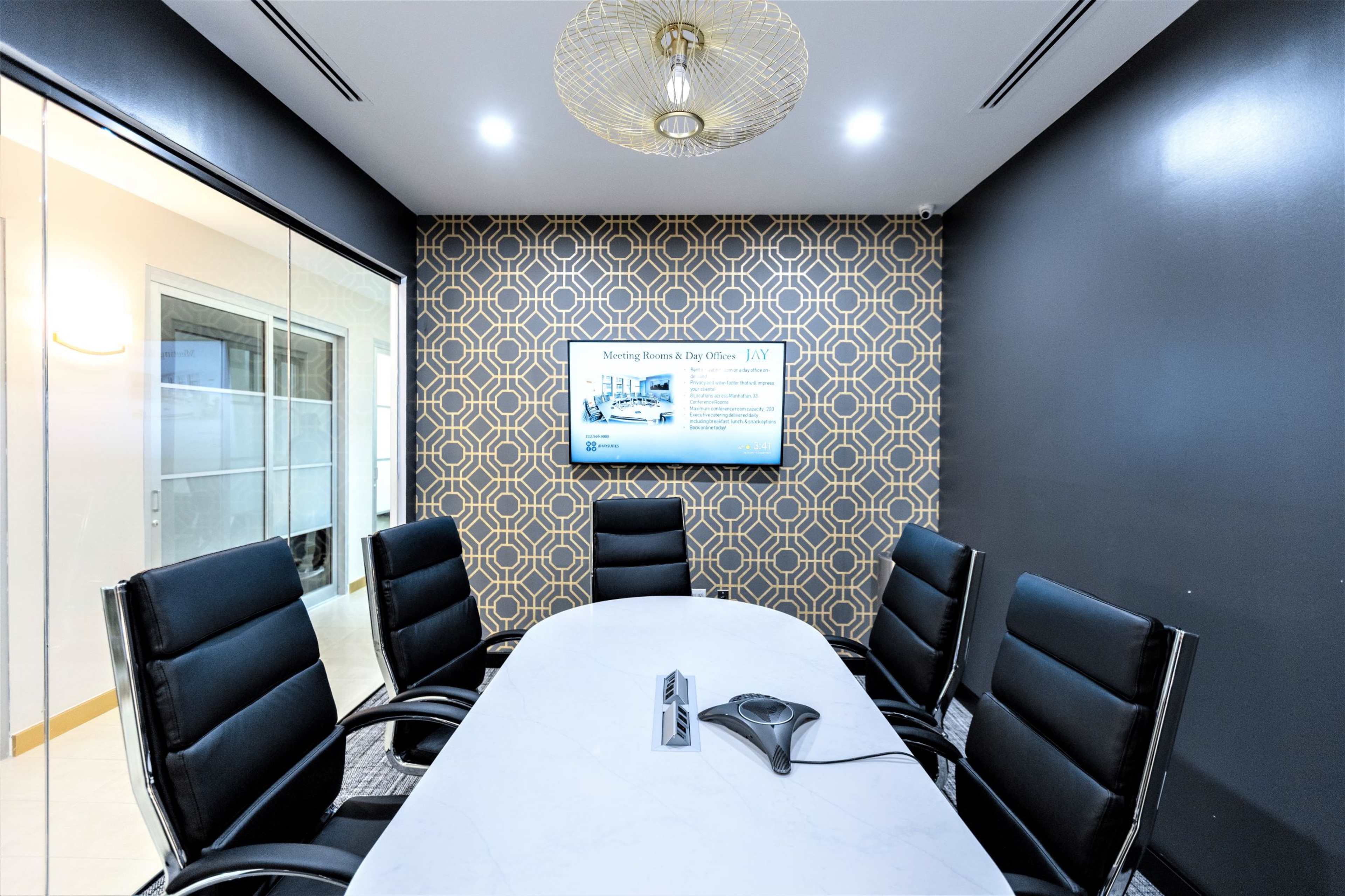 A modern conference room with a large white table, black leather chairs, and a patterned wall featuring a television screen displaying information.