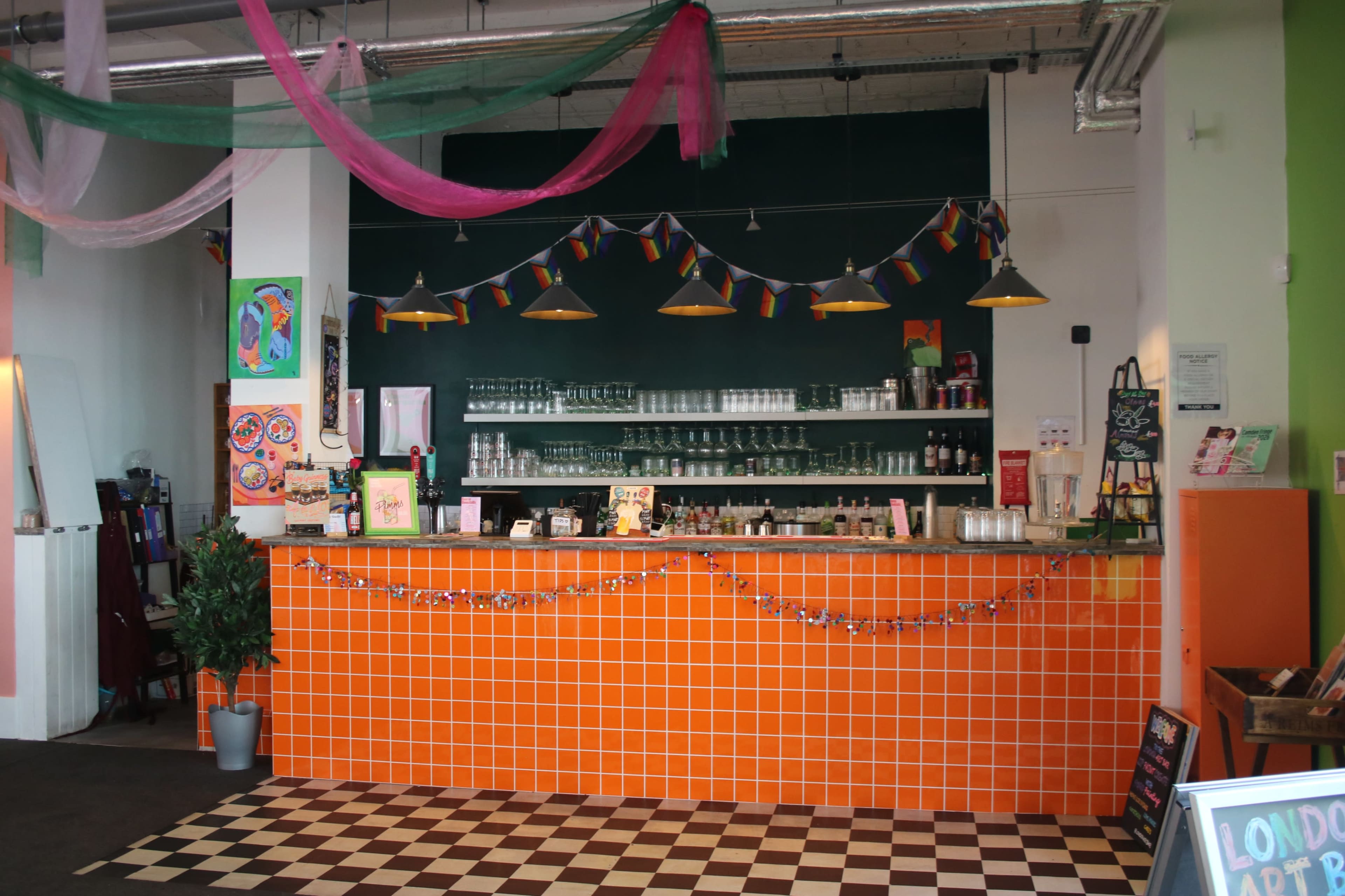 The image shows a vibrant café with an orange tiled counter, glass shelves, and colorful decorations, including hanging lights and a garland.