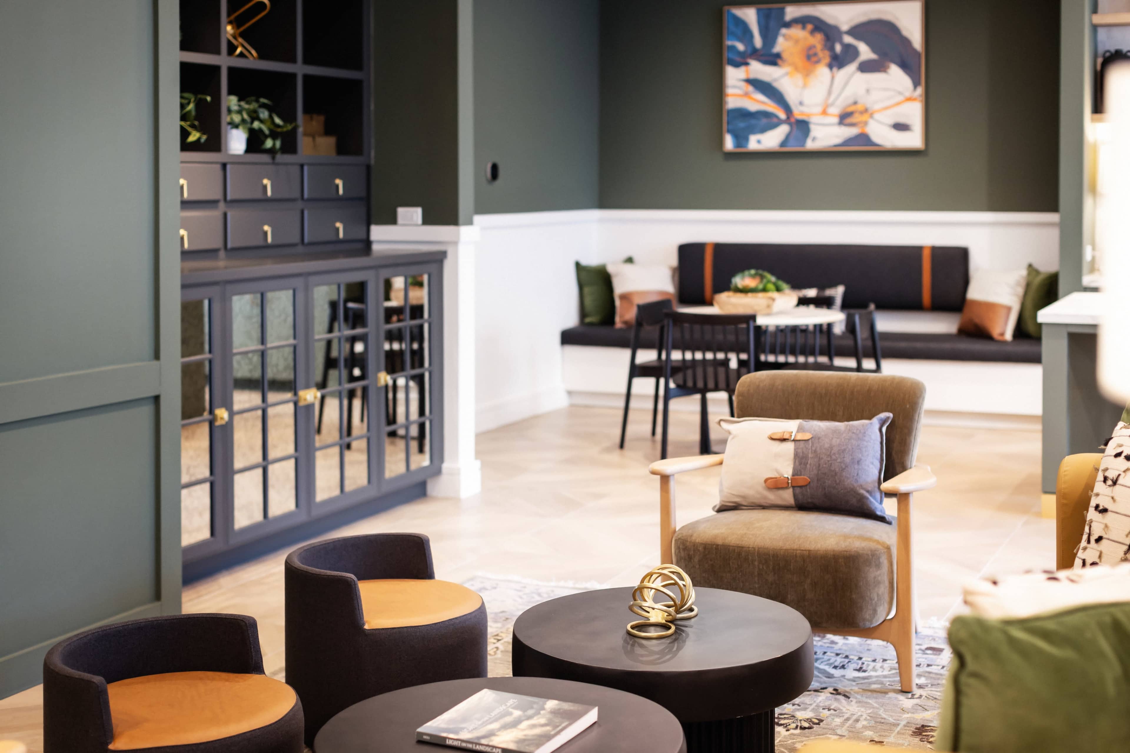 The image displays a stylish interior space featuring a cozy seating area with a combination of chairs, dark tables, and a decorative shelf, complemented by greenery and artwork on the walls.