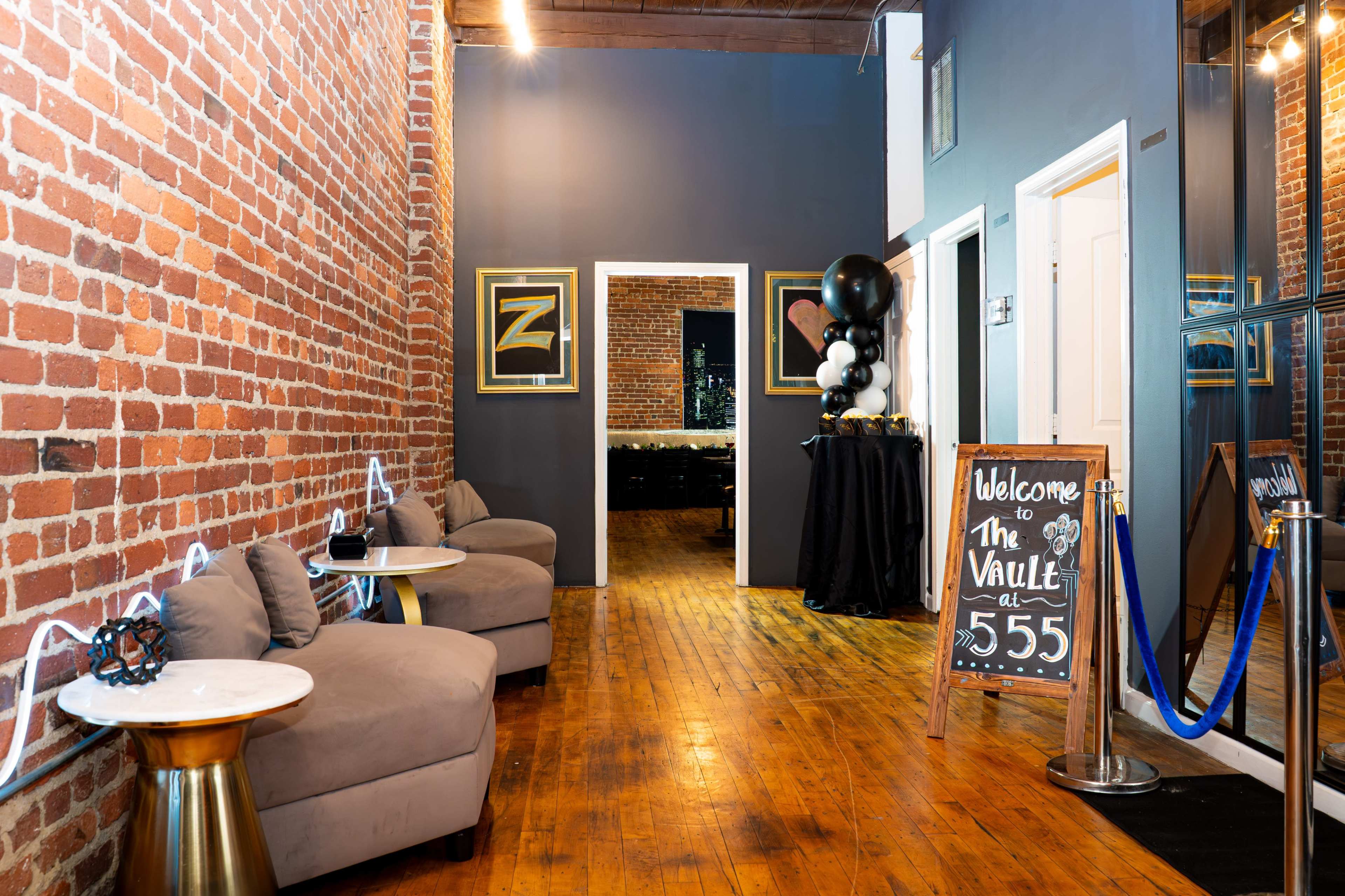 The image shows a hallway with exposed brick walls, featuring seating areas with small tables, a welcome sign, and a mirrored wall that leads to a darkened space at the end.