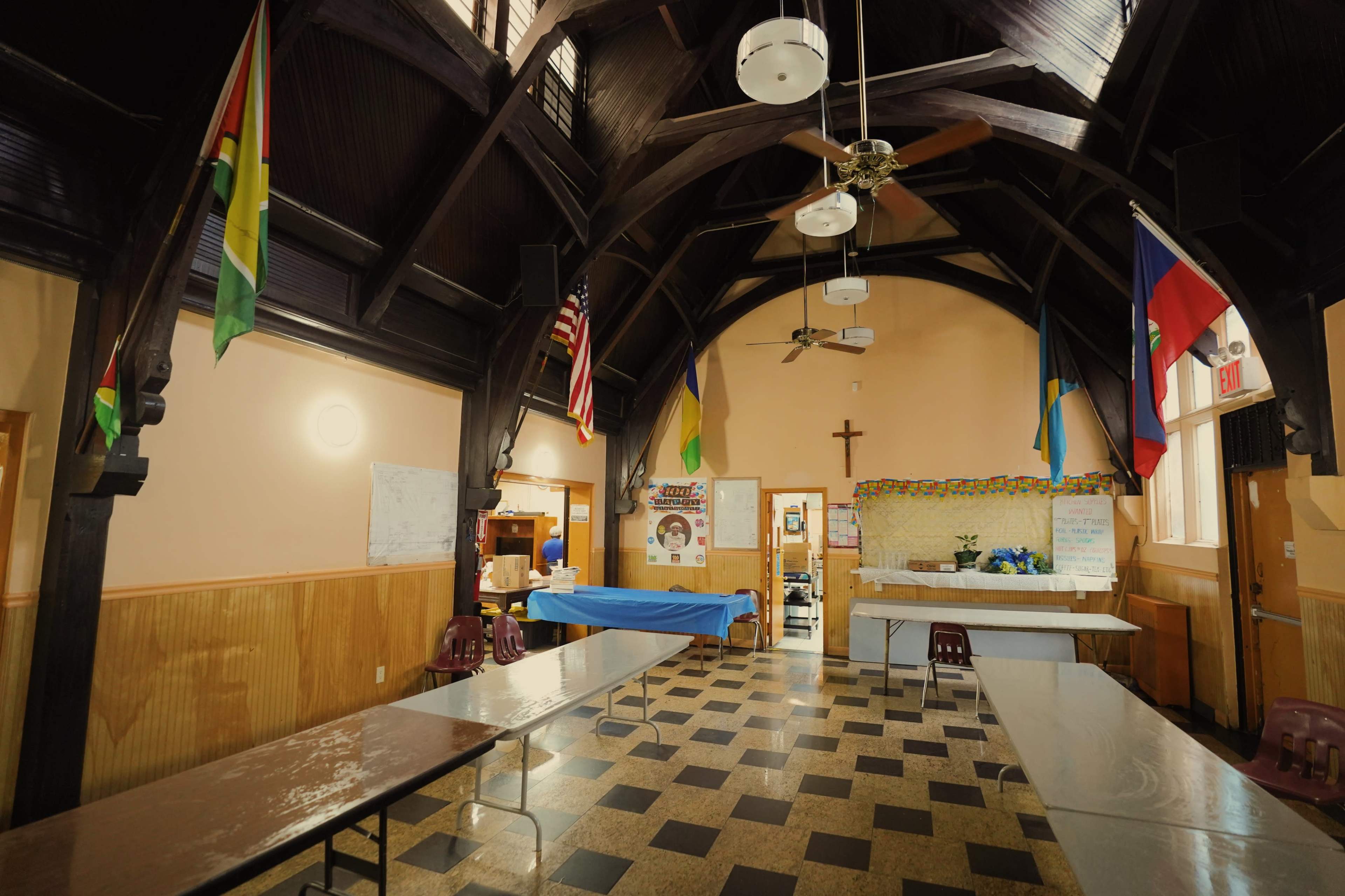 The interior of a community hall features a high, wooden-beamed ceiling, several tables, and various flags hanging on the walls.
