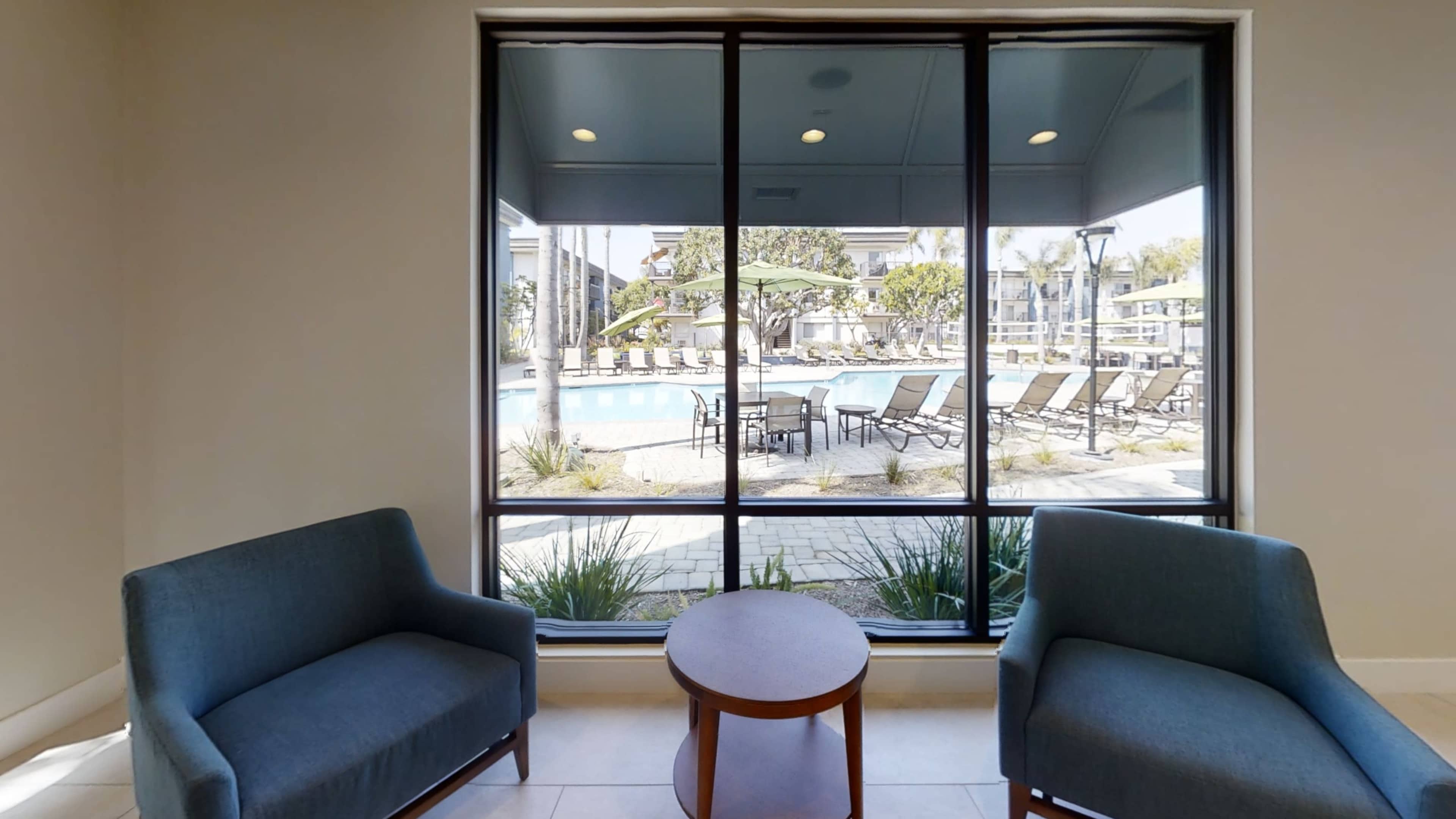 Two blue chairs and a small round table are positioned in front of large windows overlooking a pool area with lounge chairs and greenery.