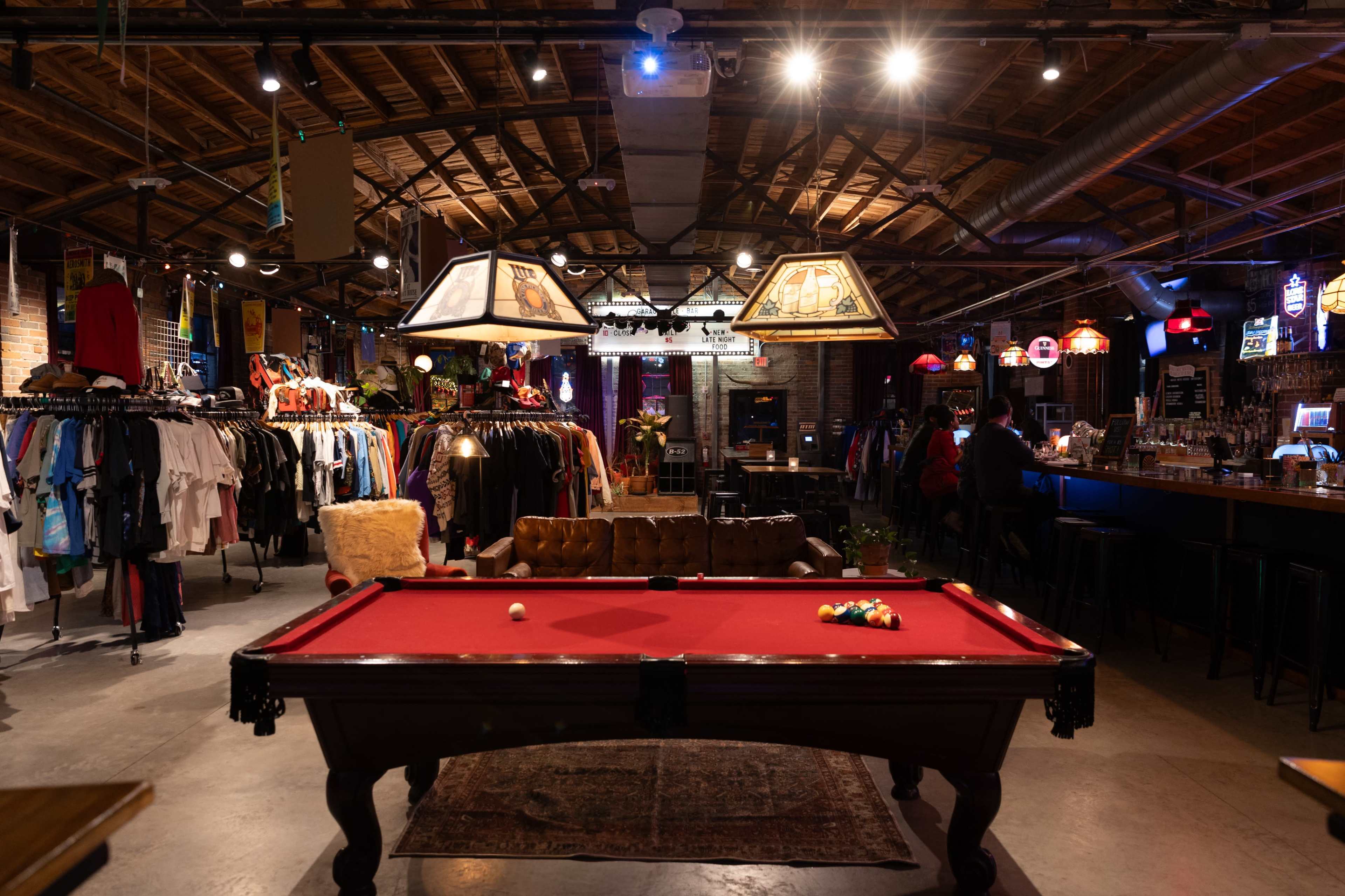 The image shows a vibrant bar and retail space featuring a pool table in the center, surrounded by clothing racks and lounge seating under warm lighting.