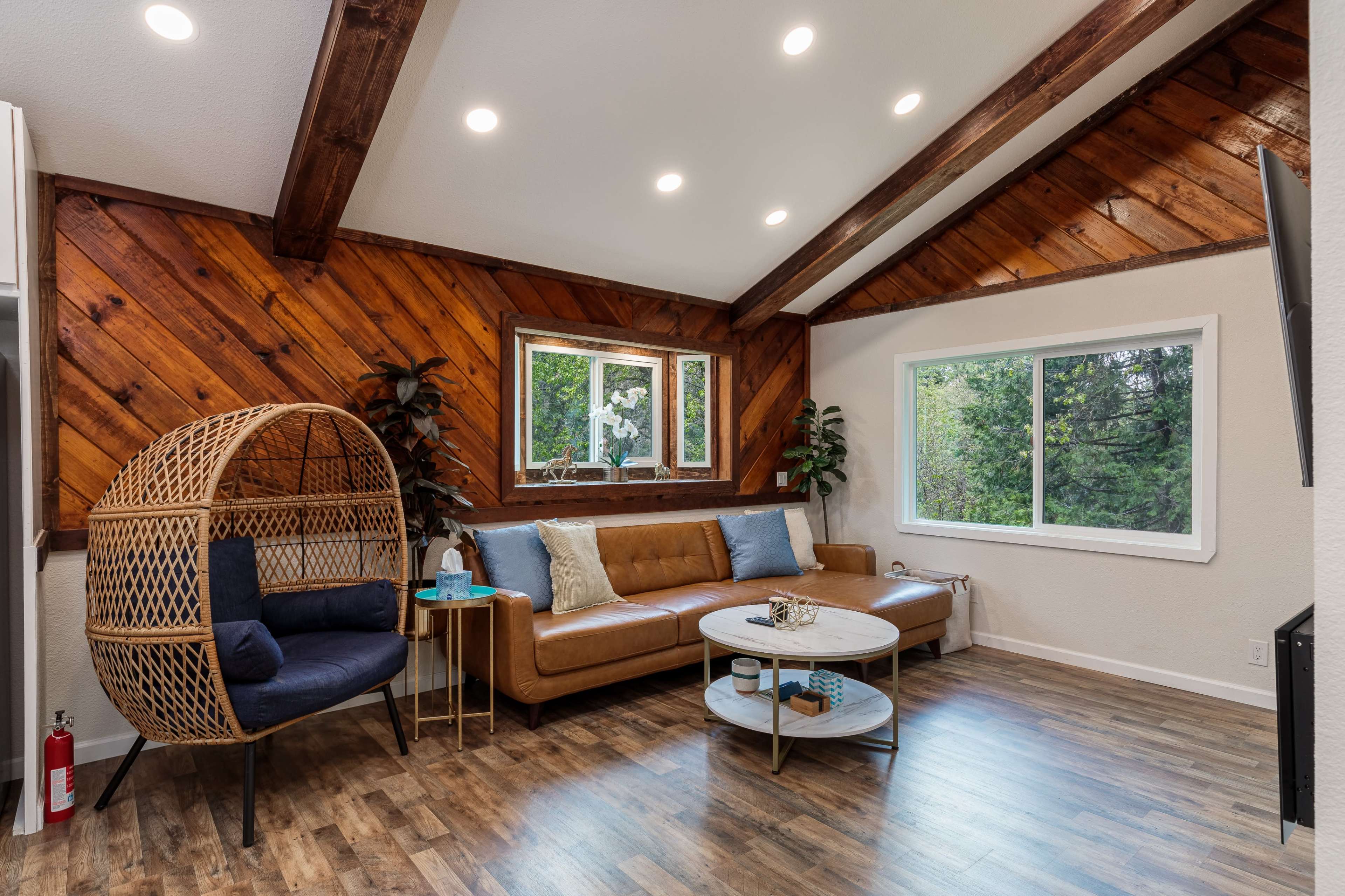 A cozy living room featuring a brown leather sofa, a round coffee table, and a unique wicker chair next to a window with a view of greenery outside.