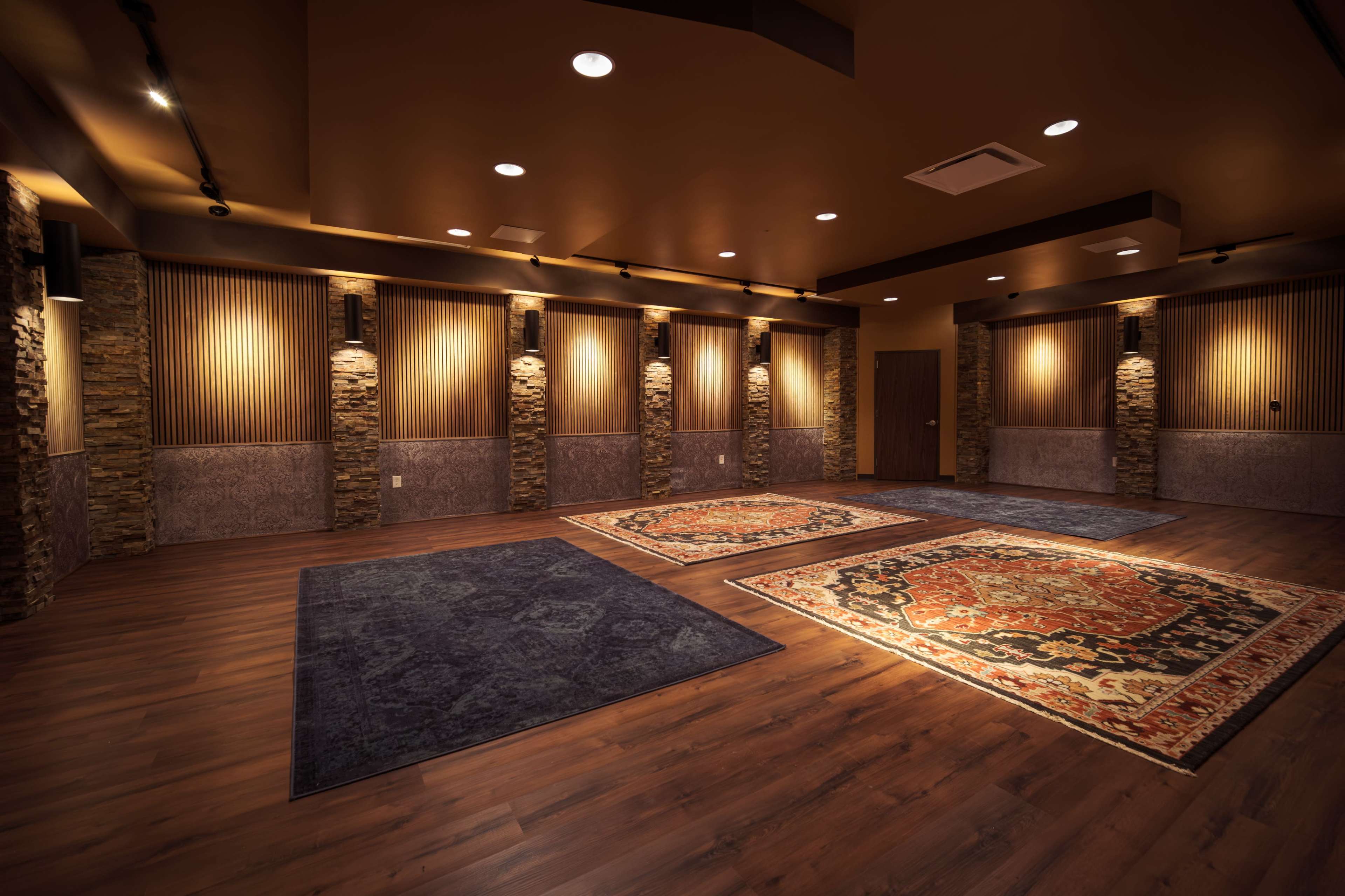 The image shows a spacious room with hardwood flooring, stone-accented walls, and area rugs placed on the floor.