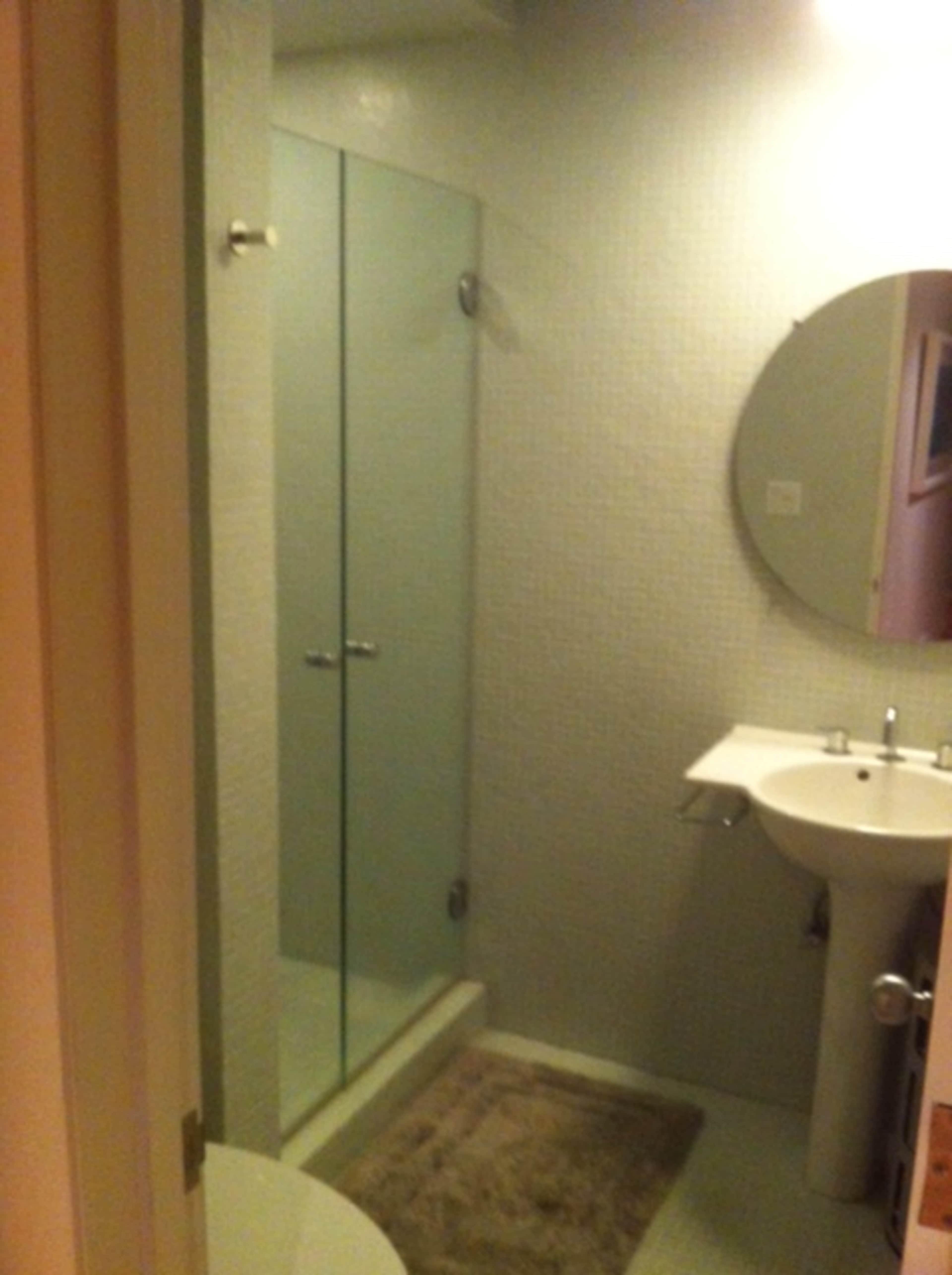 The image shows a modern bathroom with a glass shower enclosure, a round mirror over a sink, and a towel on the floor.