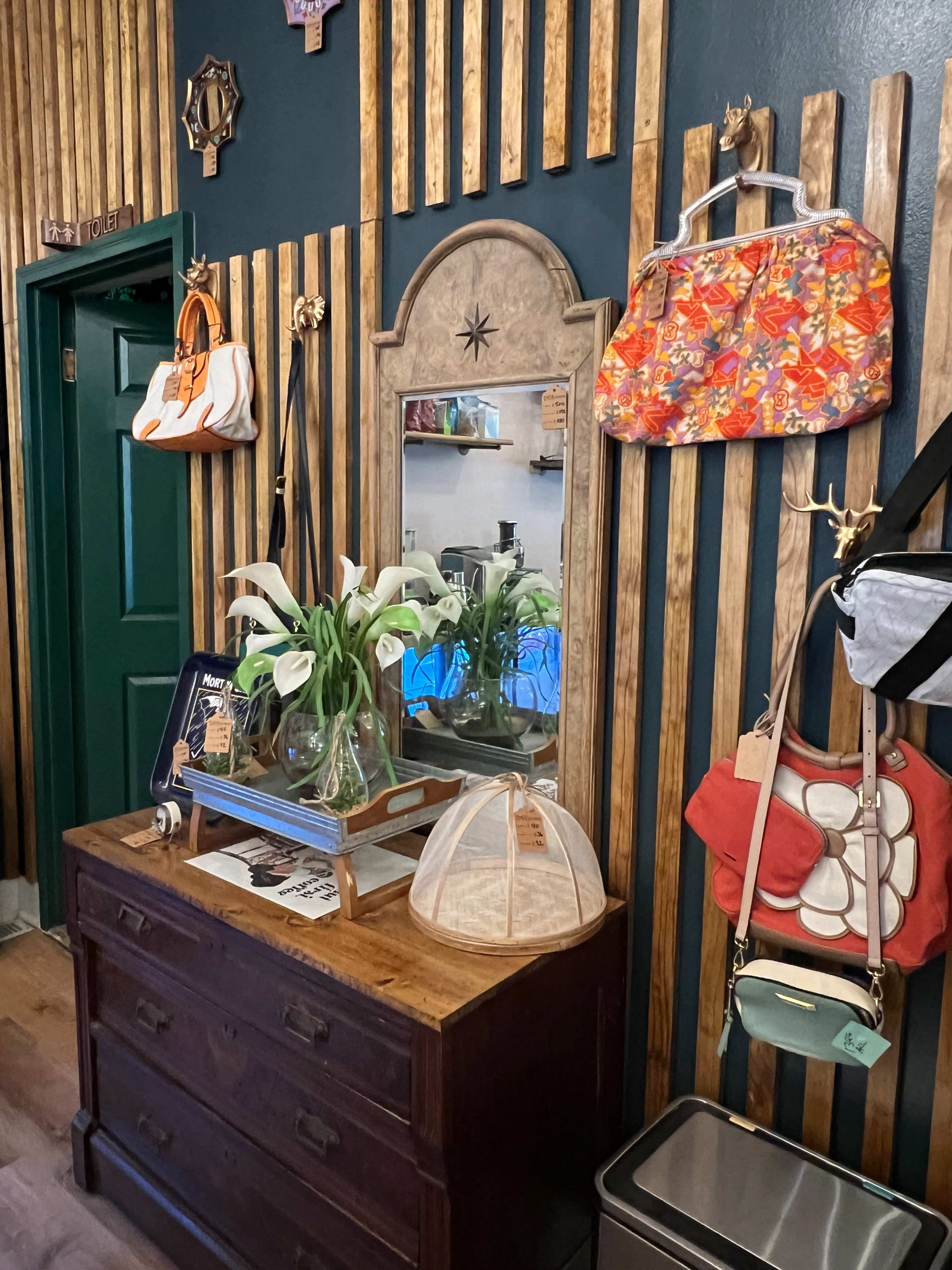 A wooden dresser against a wall with vertical wooden slats, decorated with various handbags and a mirror above a vase of flowers.