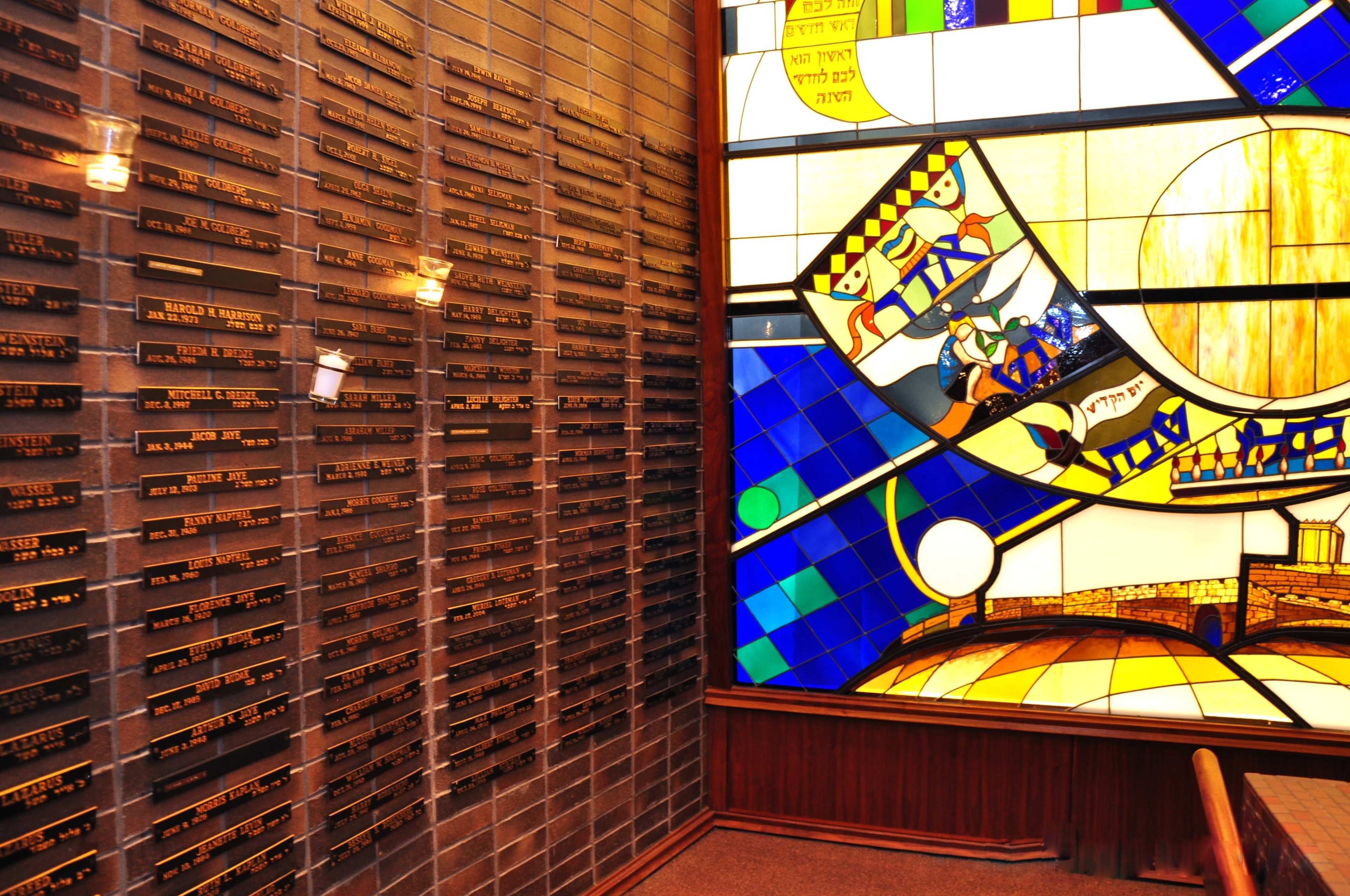 The image shows a wall with plaques bearing names next to a large stained glass window displaying colorful geometric patterns and figures.