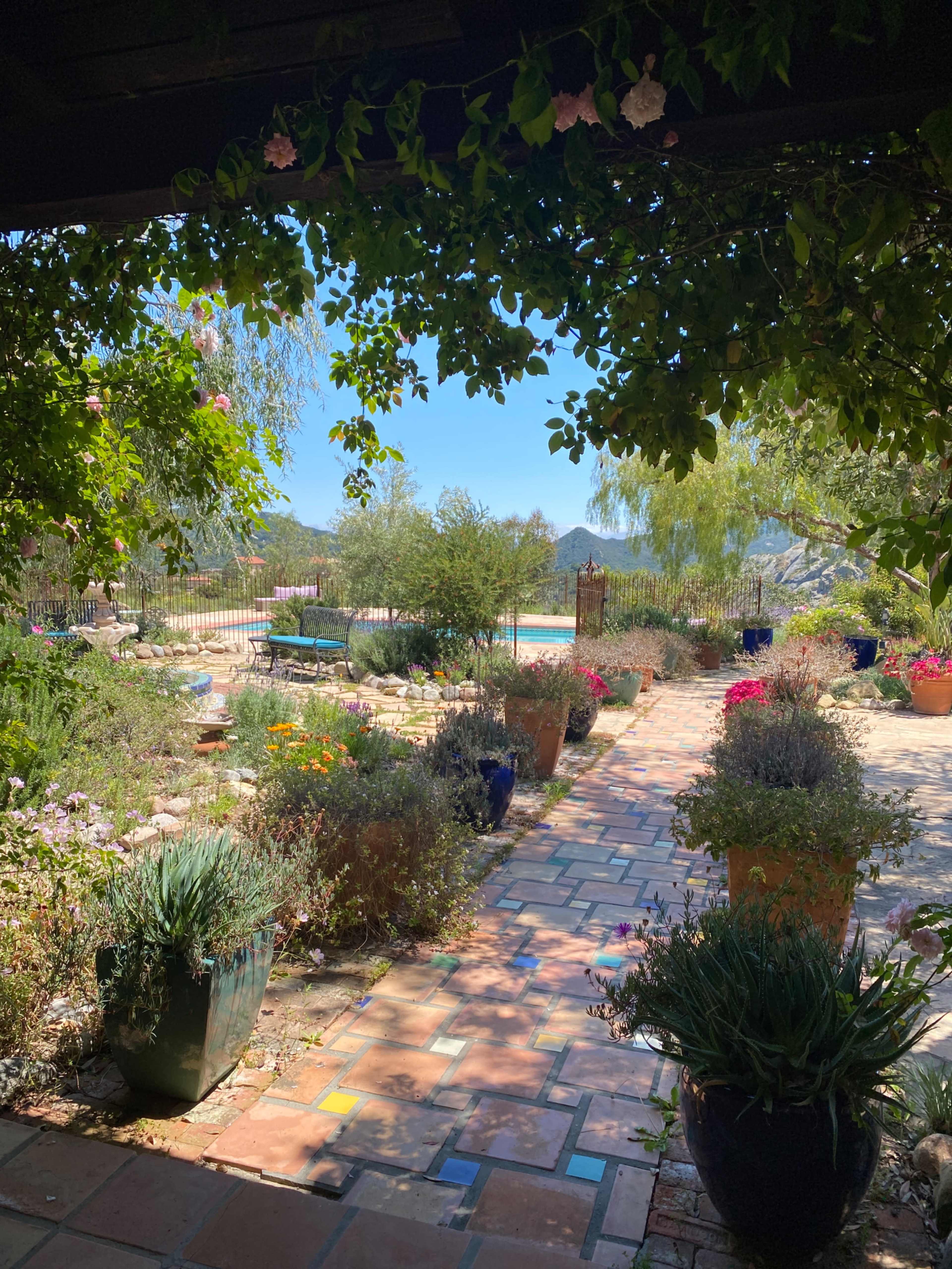 A stone pathway lined with plants and colorful flower pots leads into a garden with a view of distant hills and a blue sky.