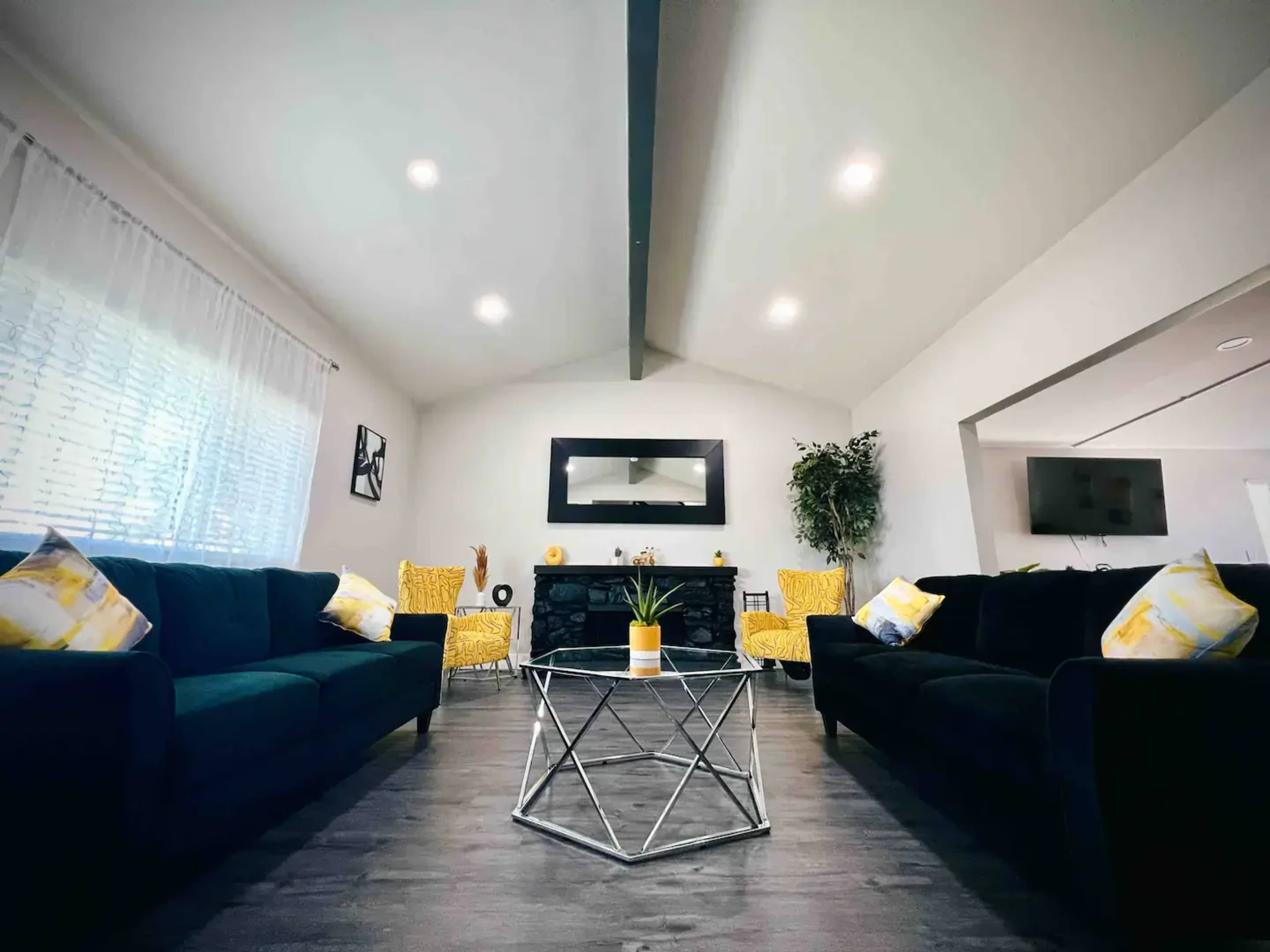A modern living room with two dark sofas, yellow accent chairs, a geometric coffee table, and a large mirror on the wall.