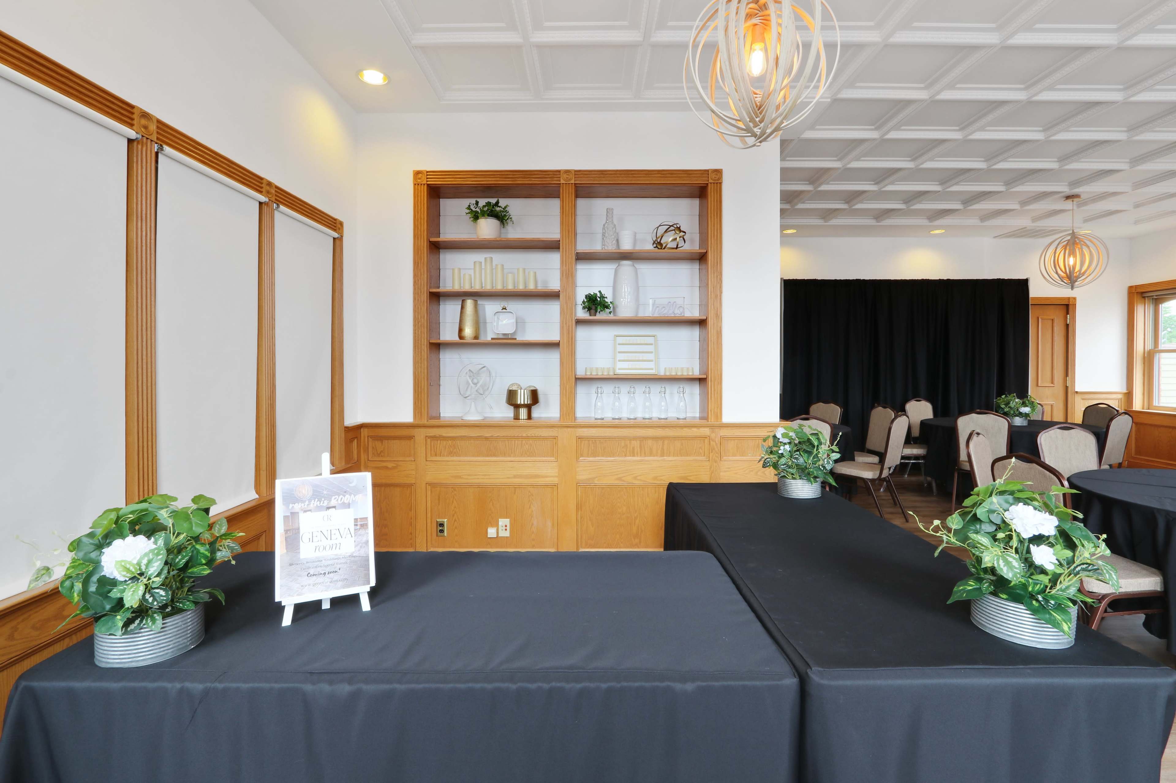 A room features a dark table with a sign in front, flanked by potted plants, and a wooden shelving unit displaying decorative items.