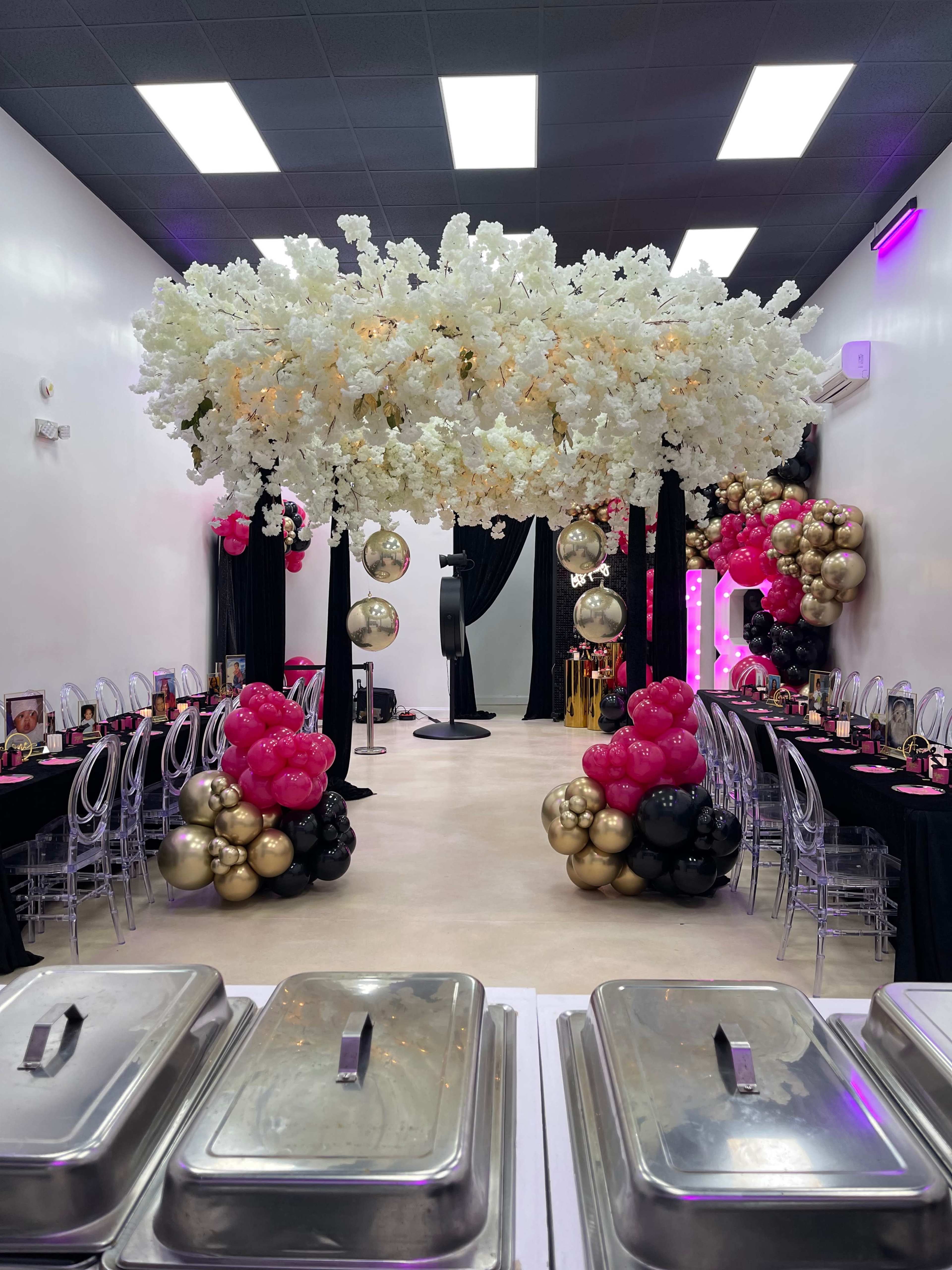 A banquet hall decorated with floral arrangements, black and pink balloon arches, and a row of buffet trays in the foreground.