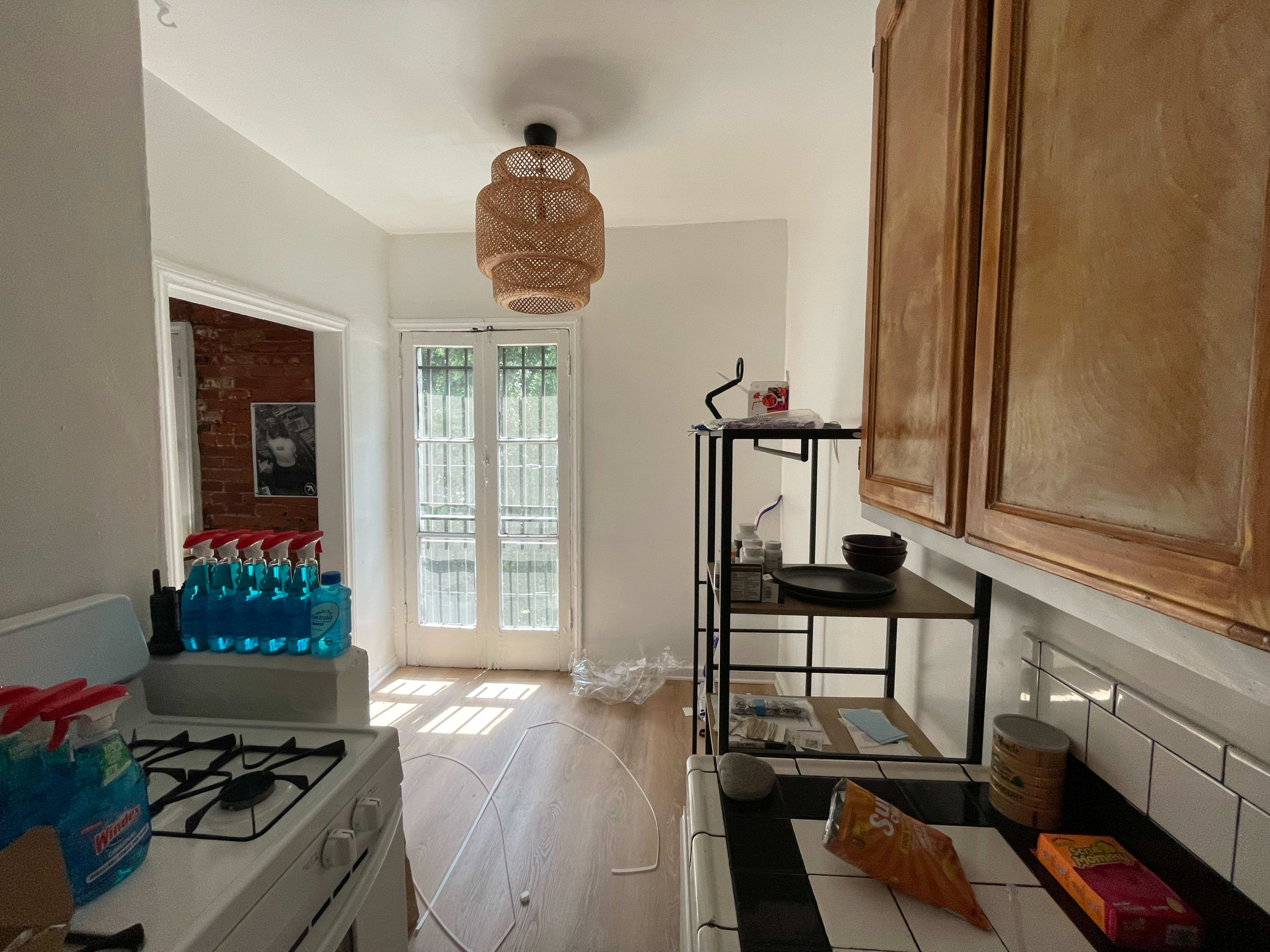 The image shows a kitchen area with a gas stove, shelves, and a doorway leading to a light-filled outdoor space, featuring a rattan light fixture and various items on surfaces.