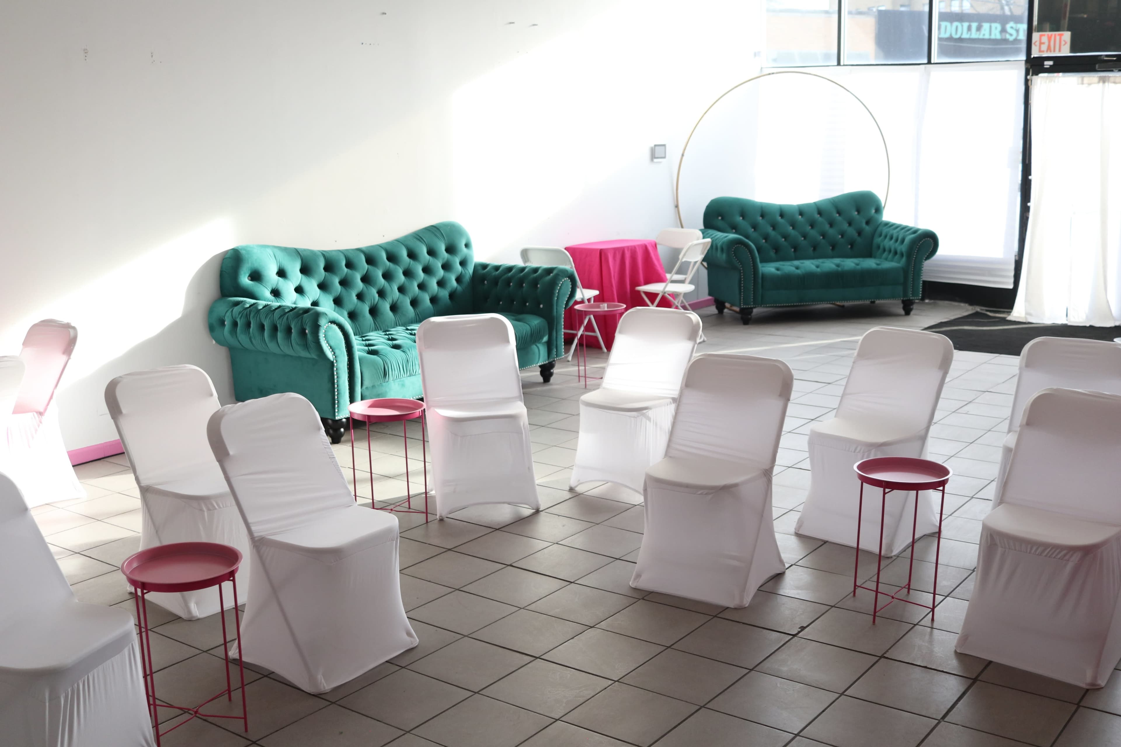The image shows a room set up for an event, featuring two green sofas, several white slipcovered chairs arranged in a circle, and pink tables in the center.