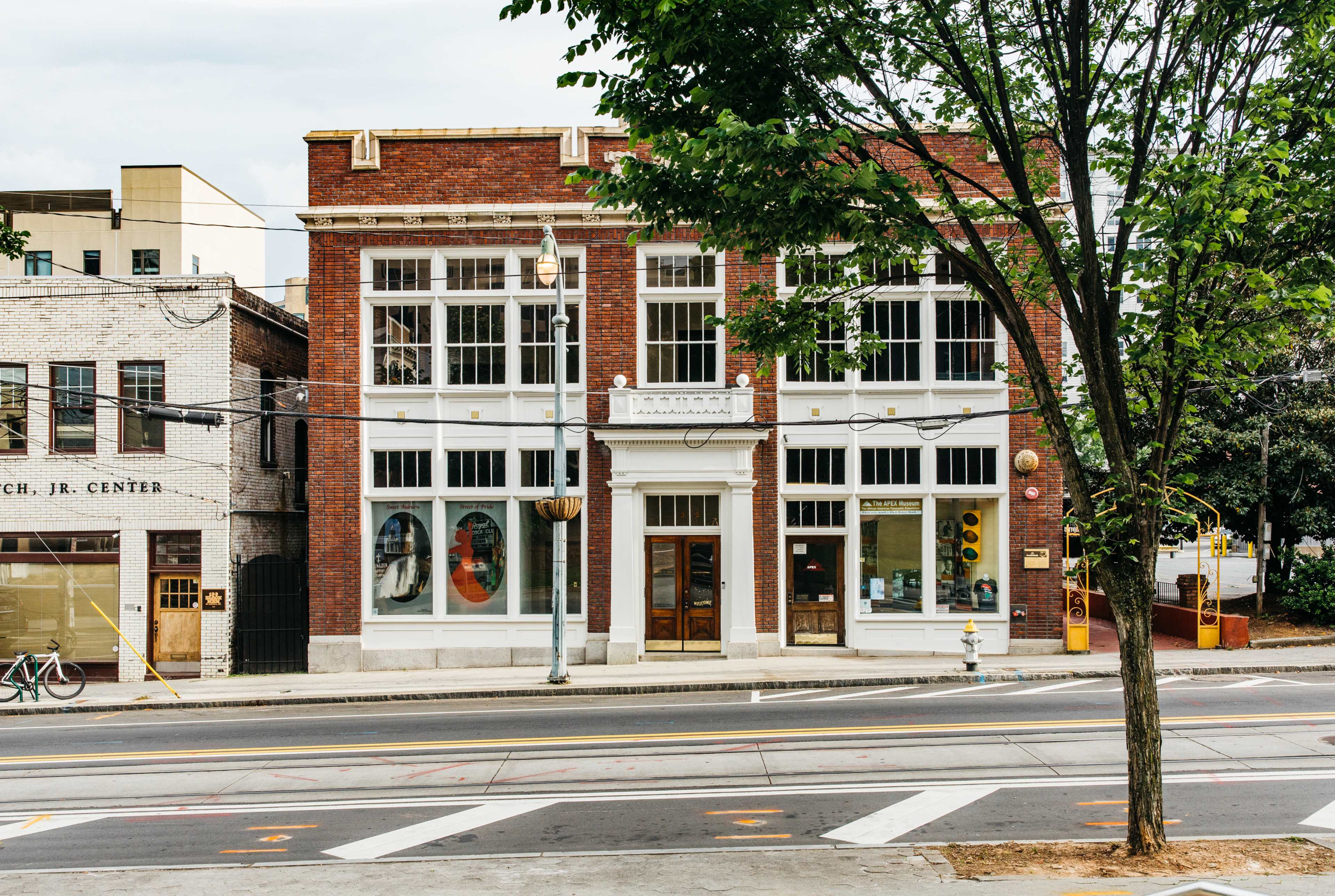 A two-story brick building with large windows stands on a street corner, surrounded by trees and a nearby bicycle.