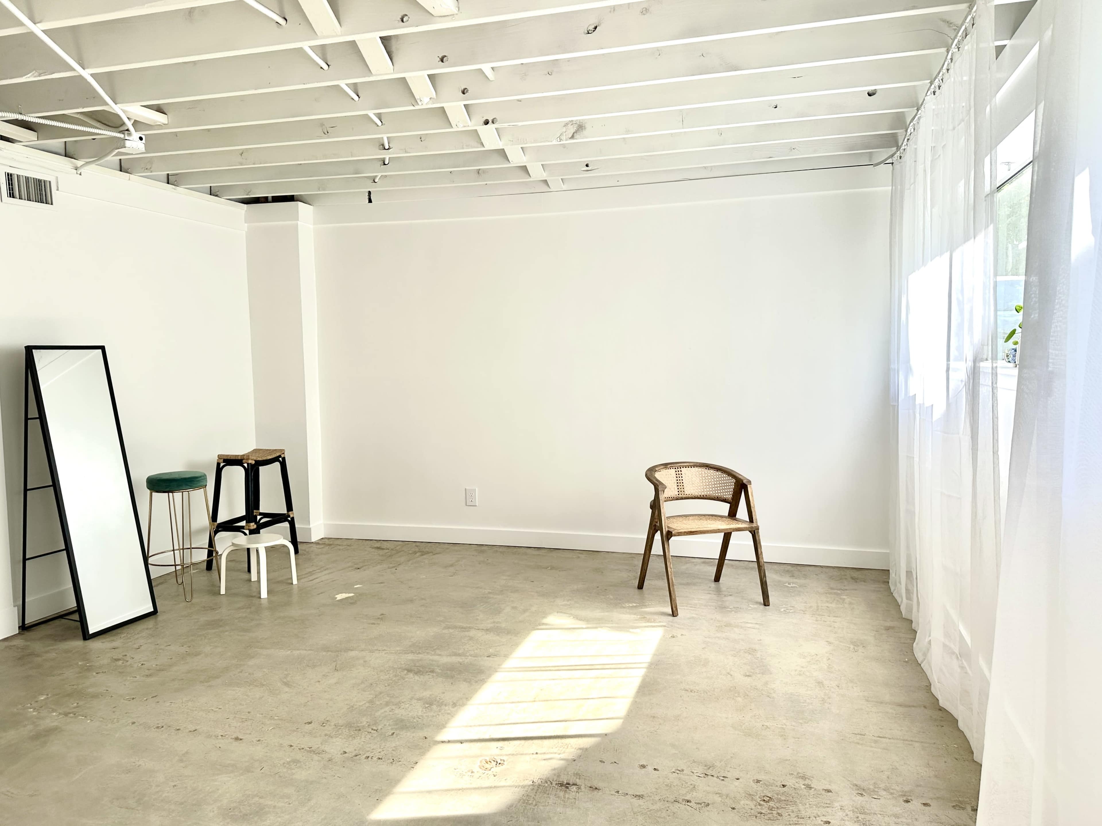 The image shows a minimalistic room with exposed ceiling beams, a mirror, a small stool, and a single chair beside a large window with sheer curtains.