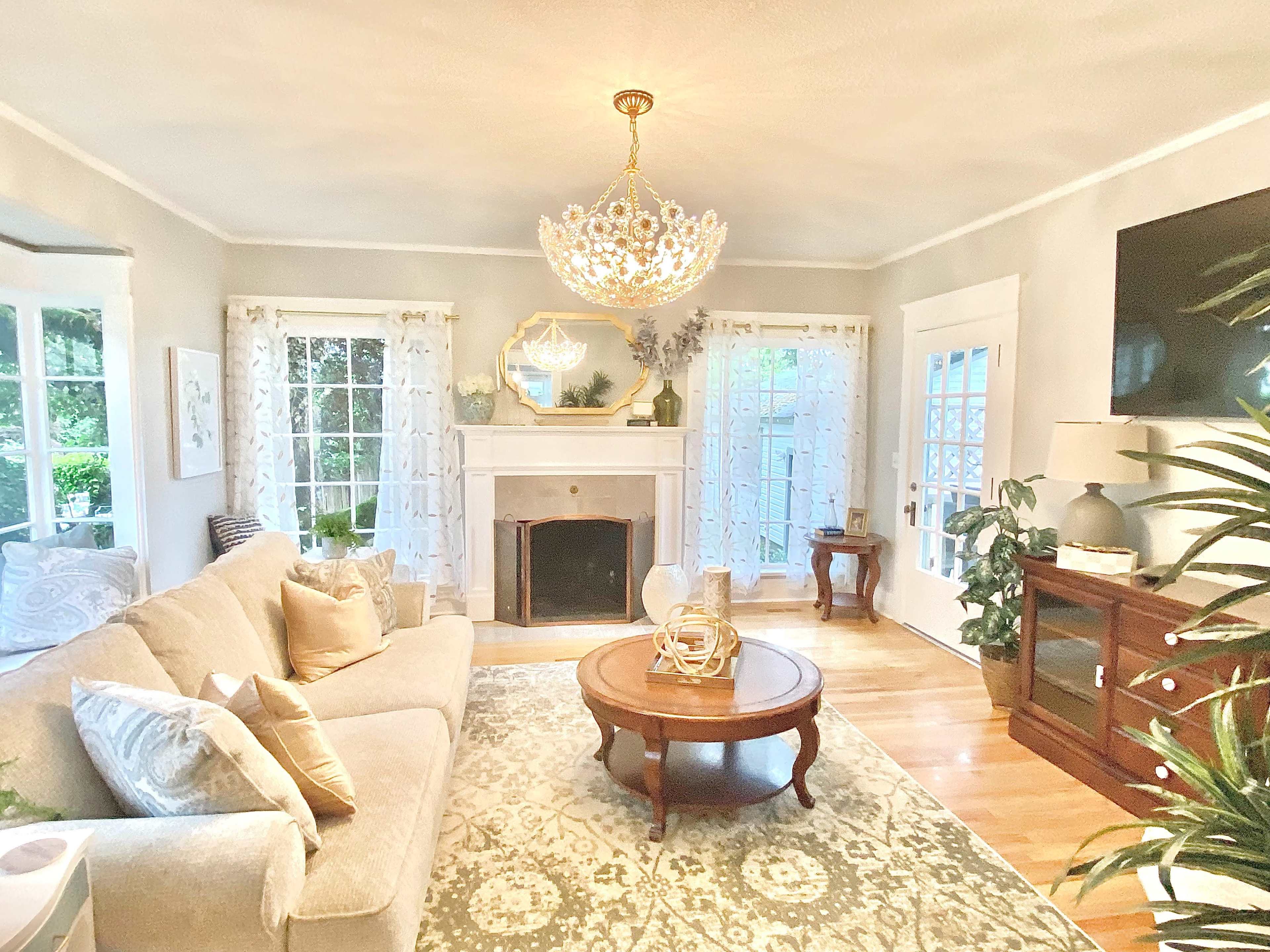 The living room features a beige sofa, a round wooden coffee table, and a large chandelier hanging from the ceiling, with large windows allowing natural light to illuminate the space.