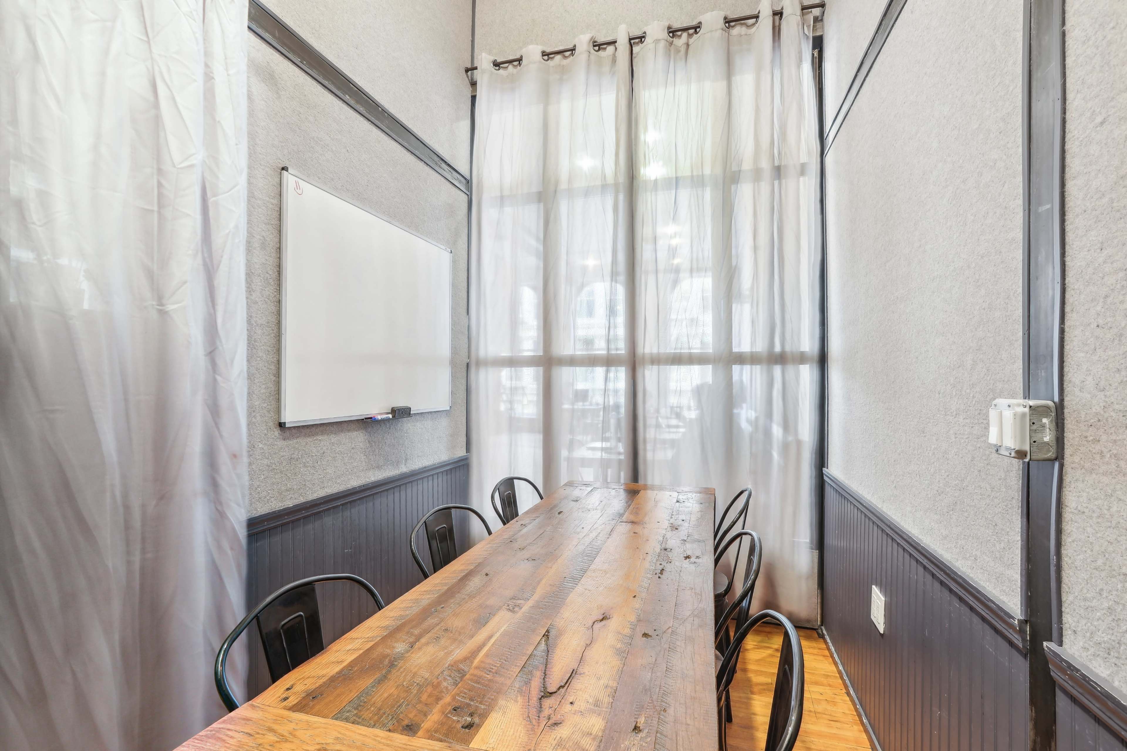 A conference room features a long wooden table surrounded by black chairs, with a whiteboard on one wall and sheer curtains allowing natural light to enter.