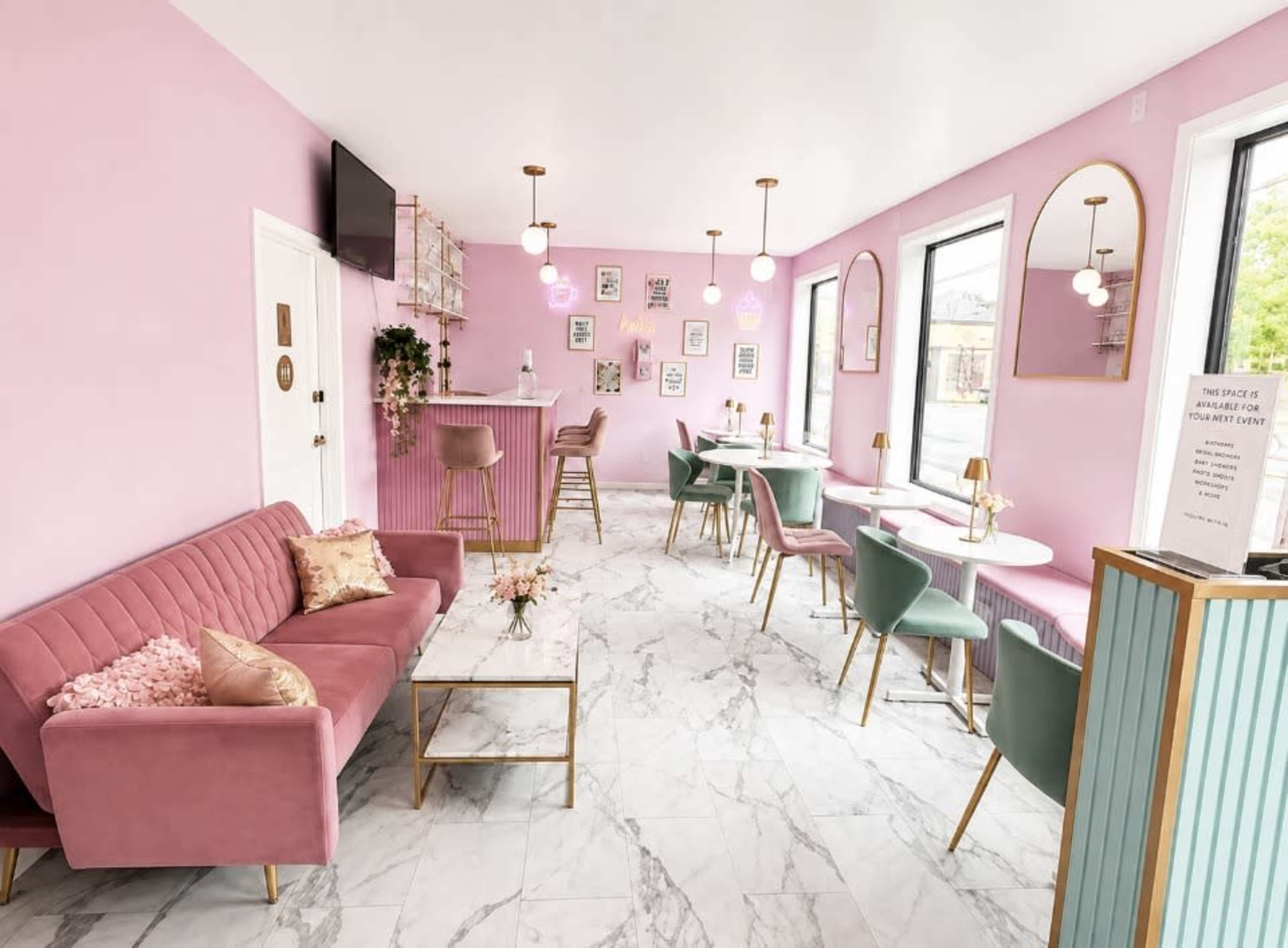 The image shows a stylish café with pink walls, marble flooring, and modern furniture including a pink sofa and green chairs arranged around small tables.