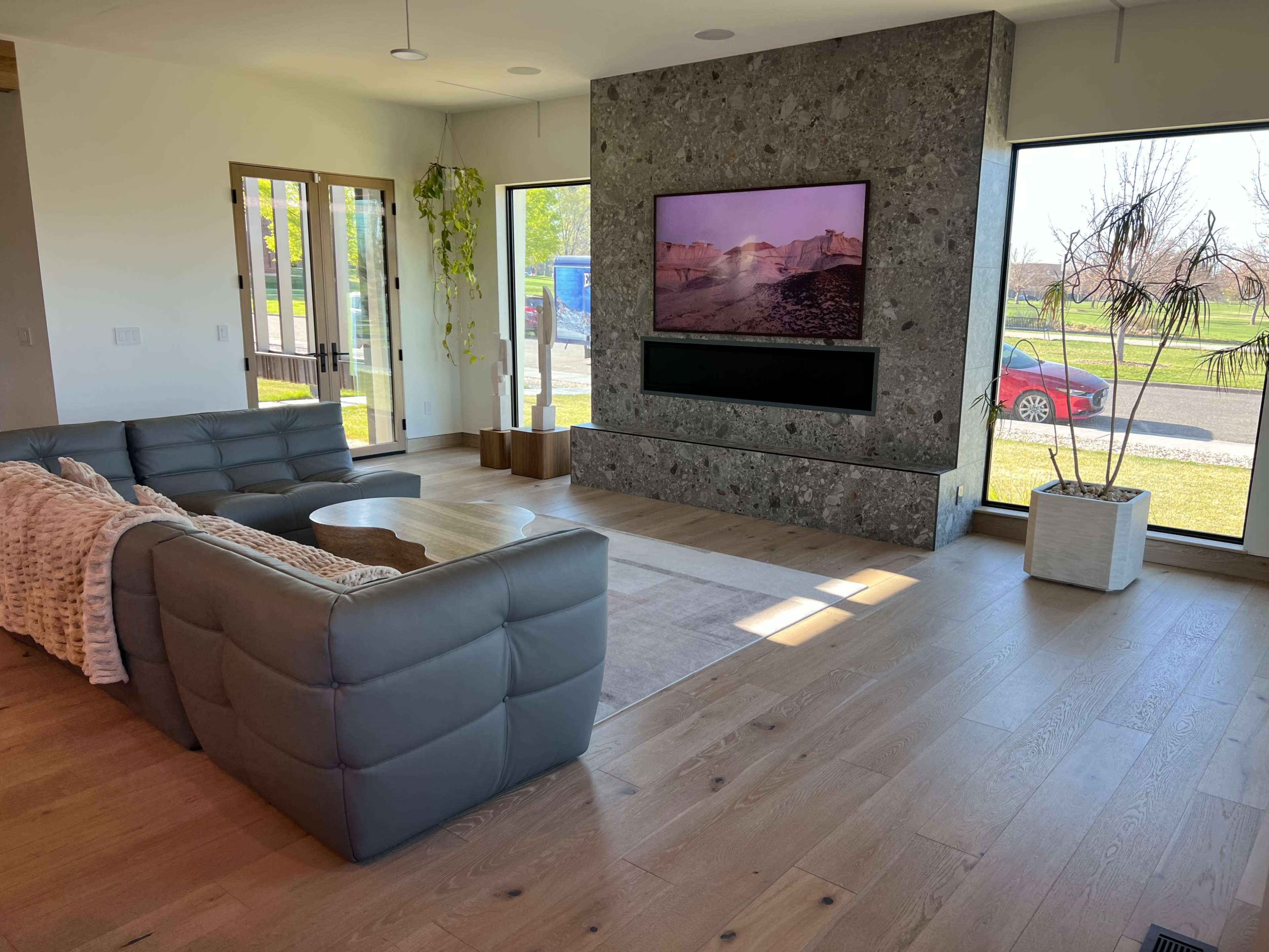 The image shows a modern living room with a gray sectional sofa, a large TV mounted on a stone wall, and large windows overlooking a garden.