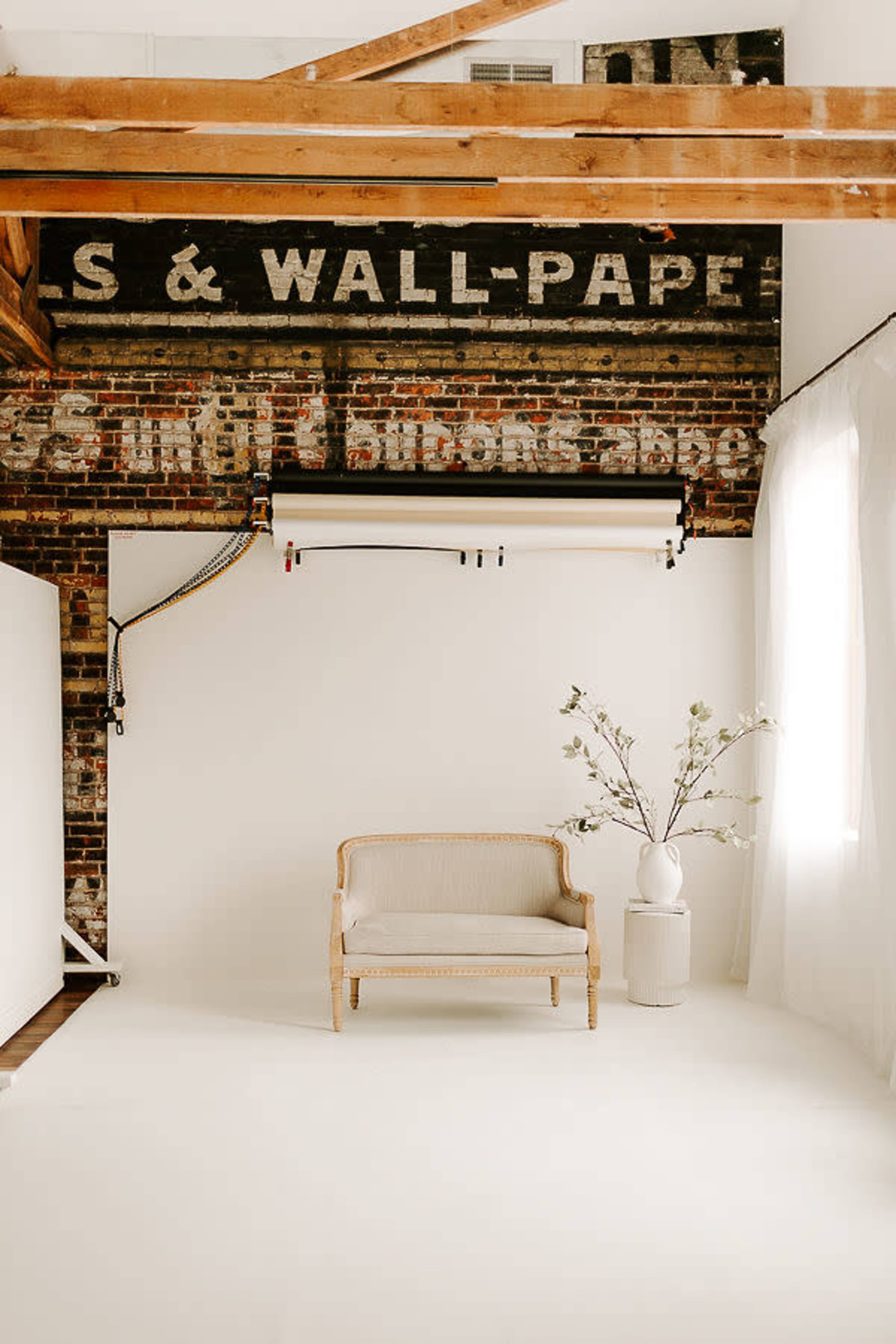 The image shows a minimalistic photo studio with a light-colored backdrop, a vintage-style sofa, and a decorative vase with flowers.