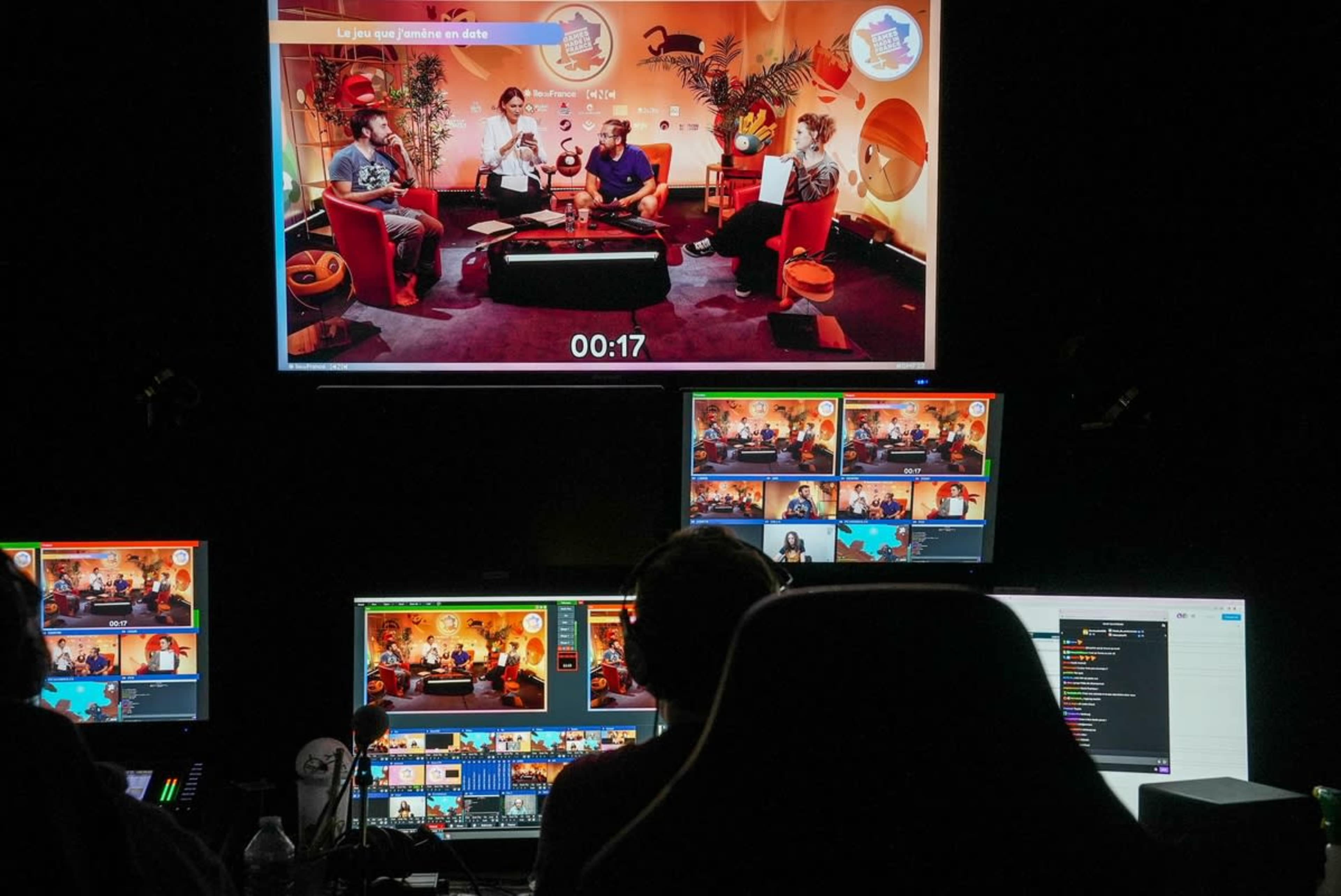 A television studio setup shows hosts discussing a topic on a large screen, with multiple monitors displaying different camera angles and production details.